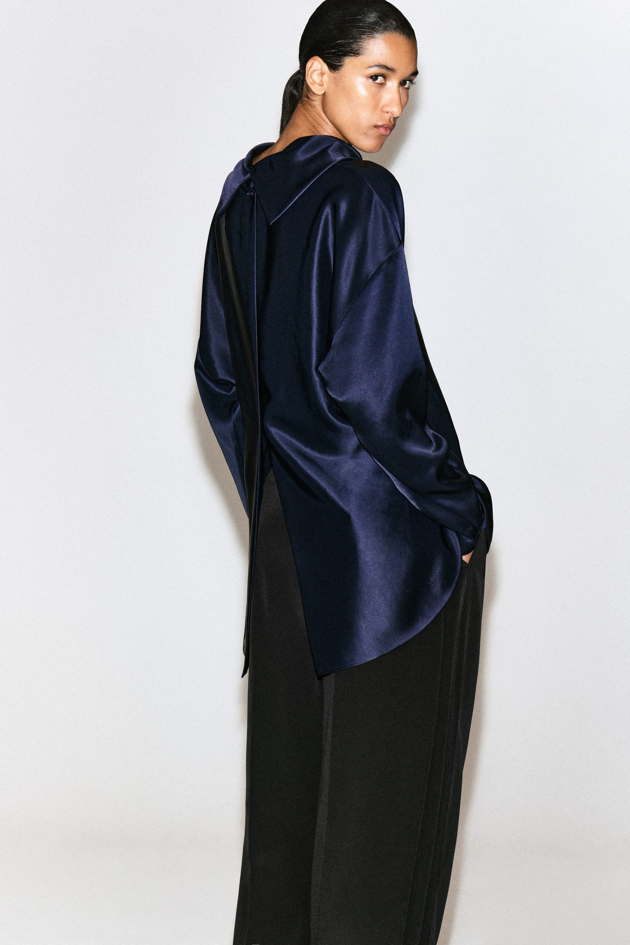 A navy blue satin blouse is shown from the back, featuring a relaxed silhouette with long sleeves and a draped, folded collar. The blouse falls over wide-leg black trousers, highlighting its split-back design and asymmetrical hem.