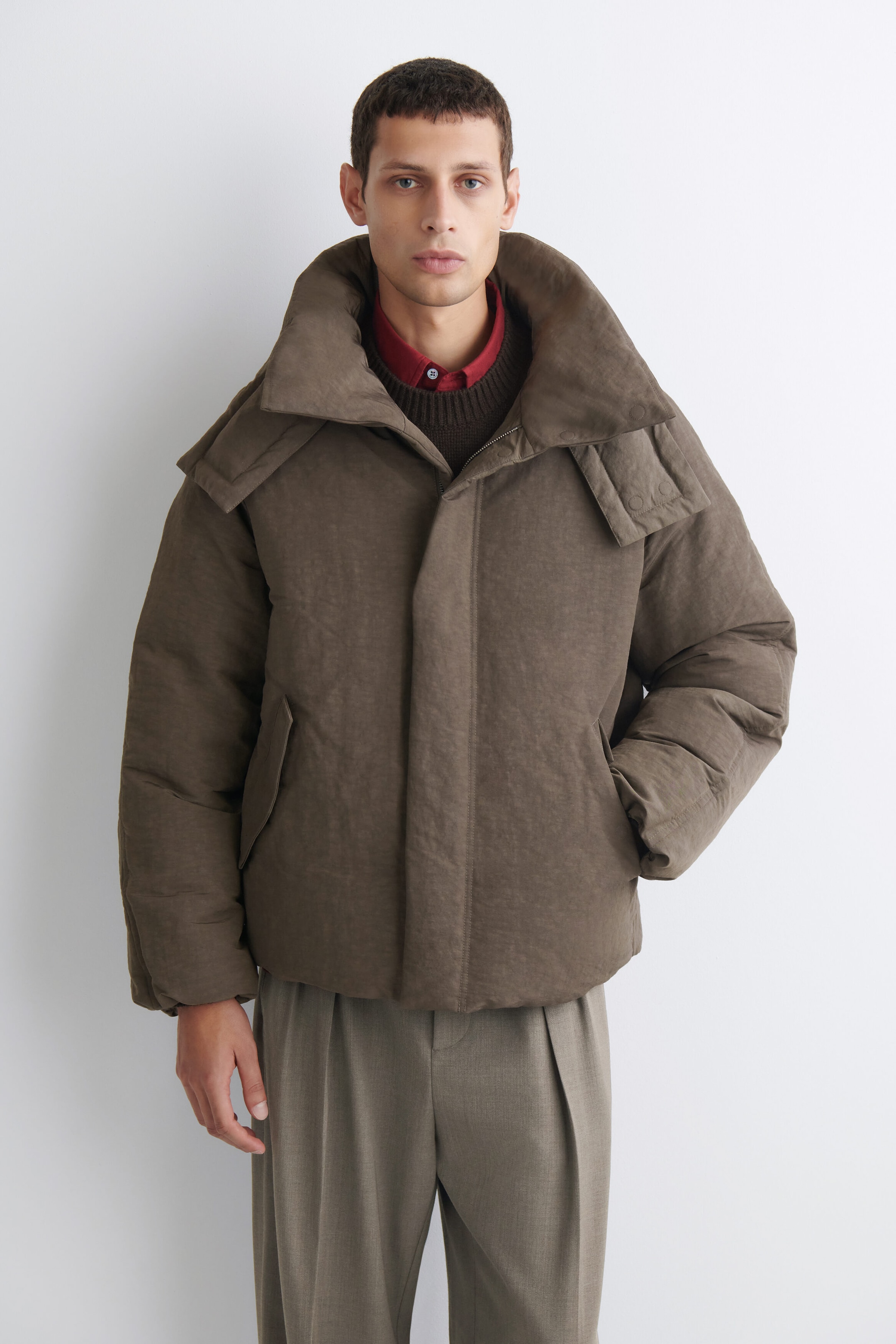 The oversized nylon-linen hooded down puffer jacket in a textured brown hue is shown from the front, featuring a voluminous high collar and concealed front placket. The jacket presents a relaxed, boxy silhouette with horizontal quilting along the body and sleeves, layered over a dark brown knit sweater and a red collared shirt. The oversized nylon-linen hooded down puffer jacket in a textured brown hue is shown from the front, featuring a voluminous high collar and concealed front placket. The jacket presents a relaxed, boxy silhouette with horizontal quilting along the body and sleeves, layered over a dark brown knit sweater and a red collared shirt.