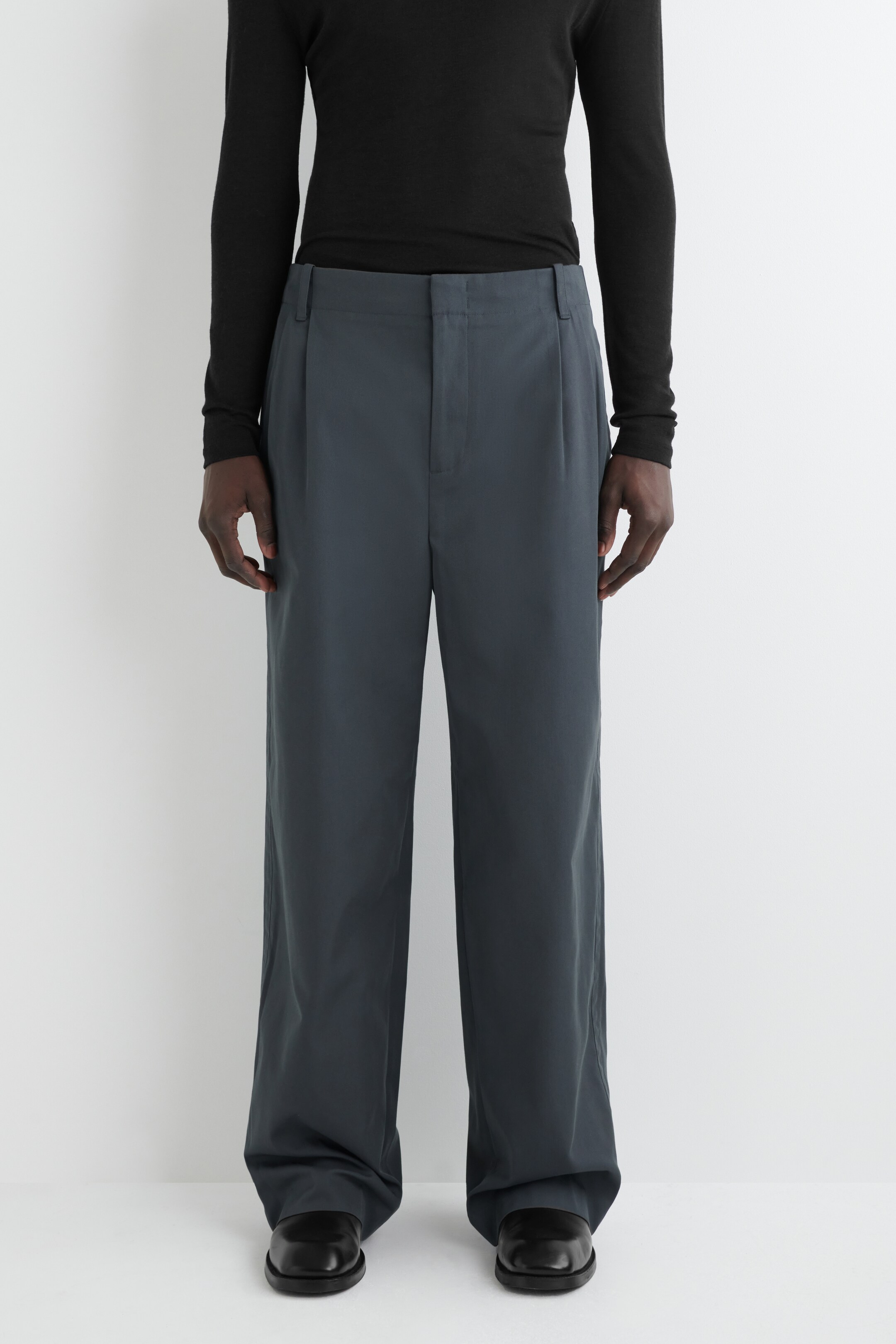 View larger image: A pair of dark grey trousers is being presented from the FRONT, featuring a relaxed, wide-leg fit. The trousers have single pleats on each leg and a concealed front closure, showcasing a smooth fabric texture.