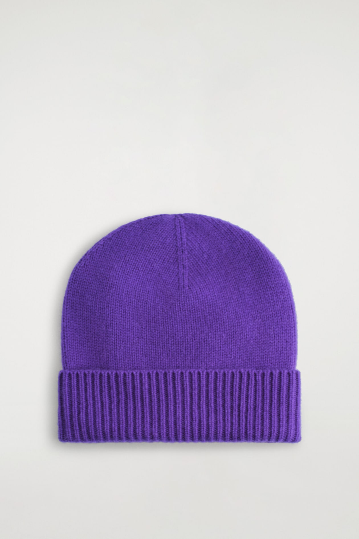 A purple cashmere beanie is shown from the front, highlighting its fine-knit texture and wide ribbed fold-over cuff. The beanie features a rounded crown with a subtle vertical seam detail at the top center.