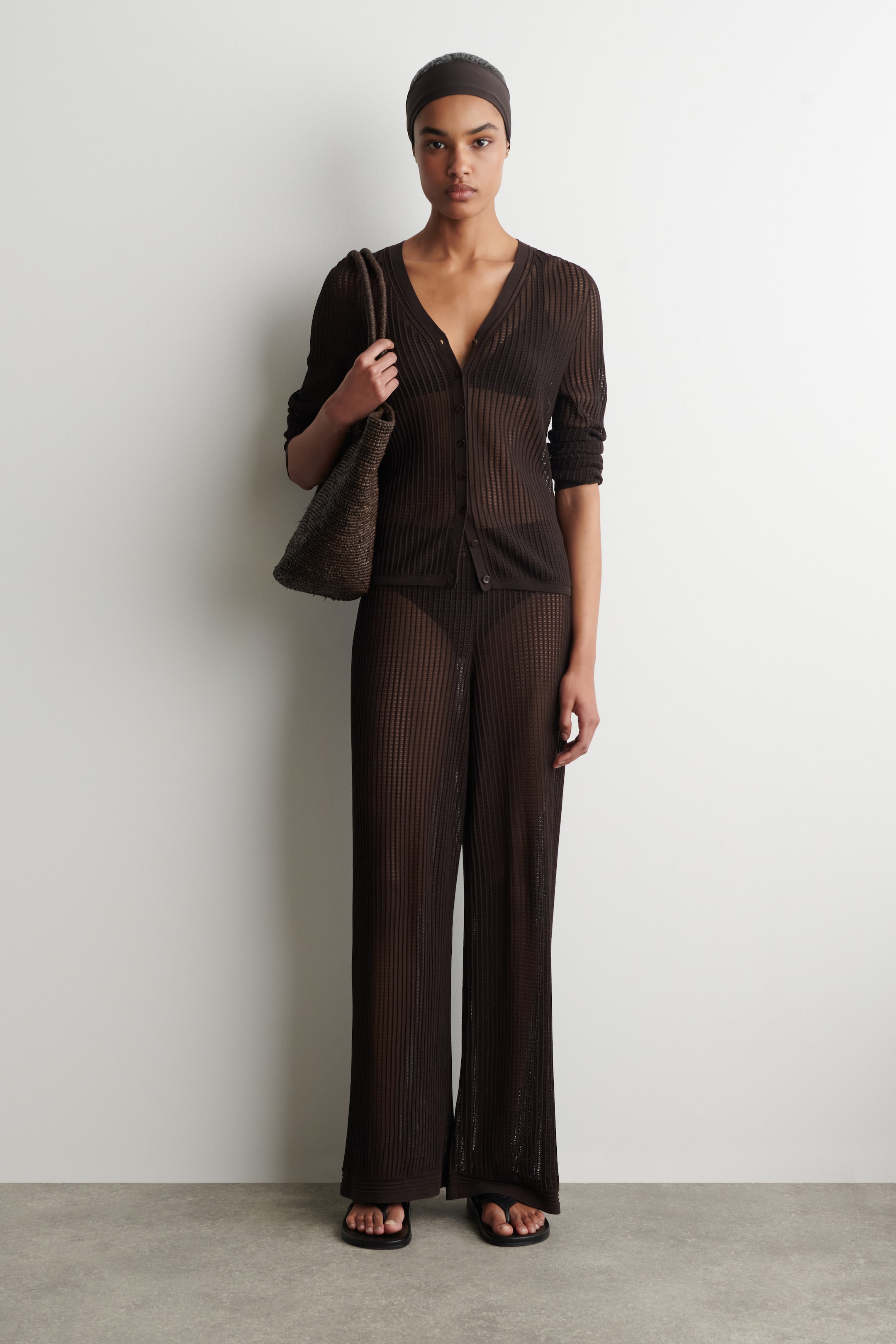 Vezi imaginea mai mare: Un cardigan cu nasturi, de culoare maro închis, din tricot cu nervuri, semi-transparent, cu decolteu în V și mâneci lungi, este purtat cu pantaloni evazați asortați. O bandă de păr simplă, de culoare maro închis, este de asemenea purtată, iar o geantă de mână împletită, de culoare maro închis, este purtată alături de sandale negre tip șlapi.