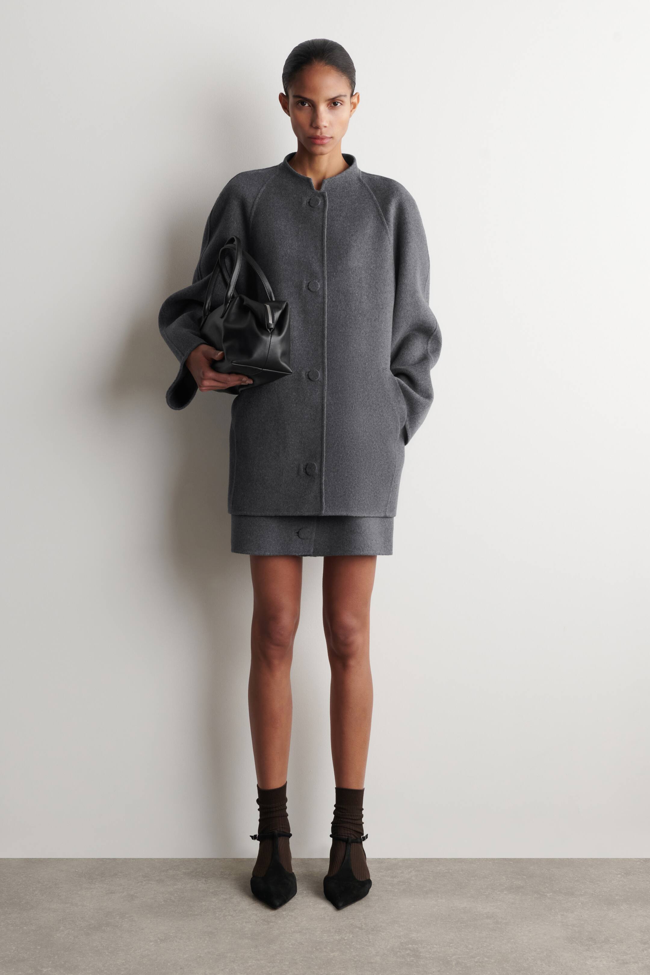 Agrandir l'image: Une veste en laine grise présente un design sans col, des manches raglan et deux boutons à l'avant, avec les mains reposant dans ses poches latérales, s'étendant jusqu'à mi-cuisse. Une mini-jupe en laine grise assortie est visible en dessous, montrant un détail de bouton unique à l'avant.