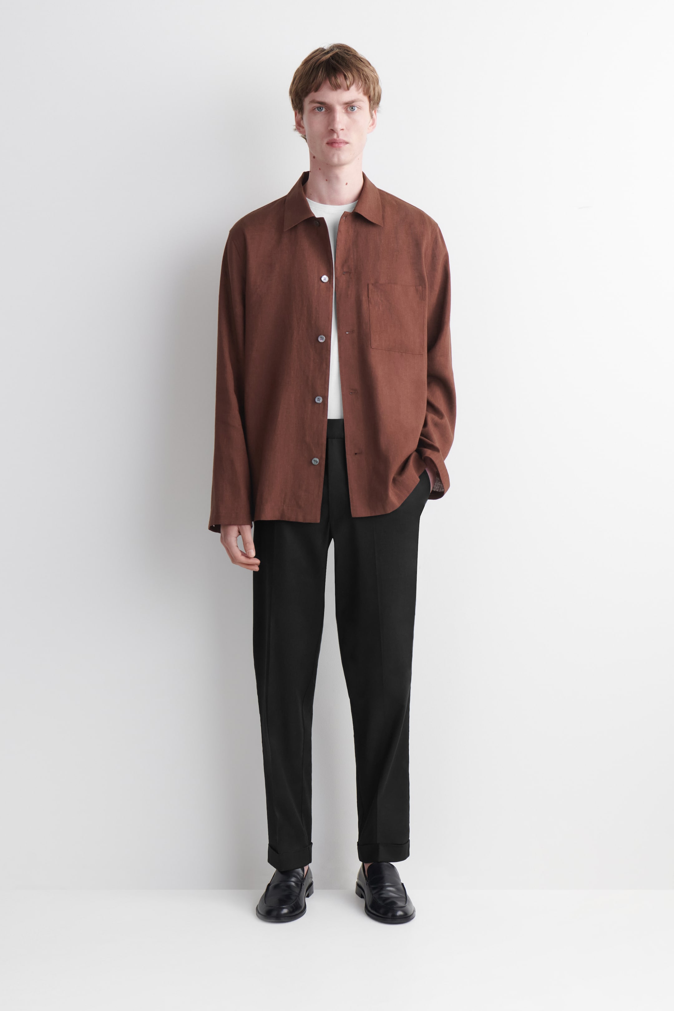 Agrandir l'image: Une chemise marron à manches longues est portée face à l'avant, déboutonnée sur un t-shirt blanc. Elle présente un col évasé, une fermeture à boutons avec des boutons foncés, et une poche plaquée unique sur la poitrine gauche.