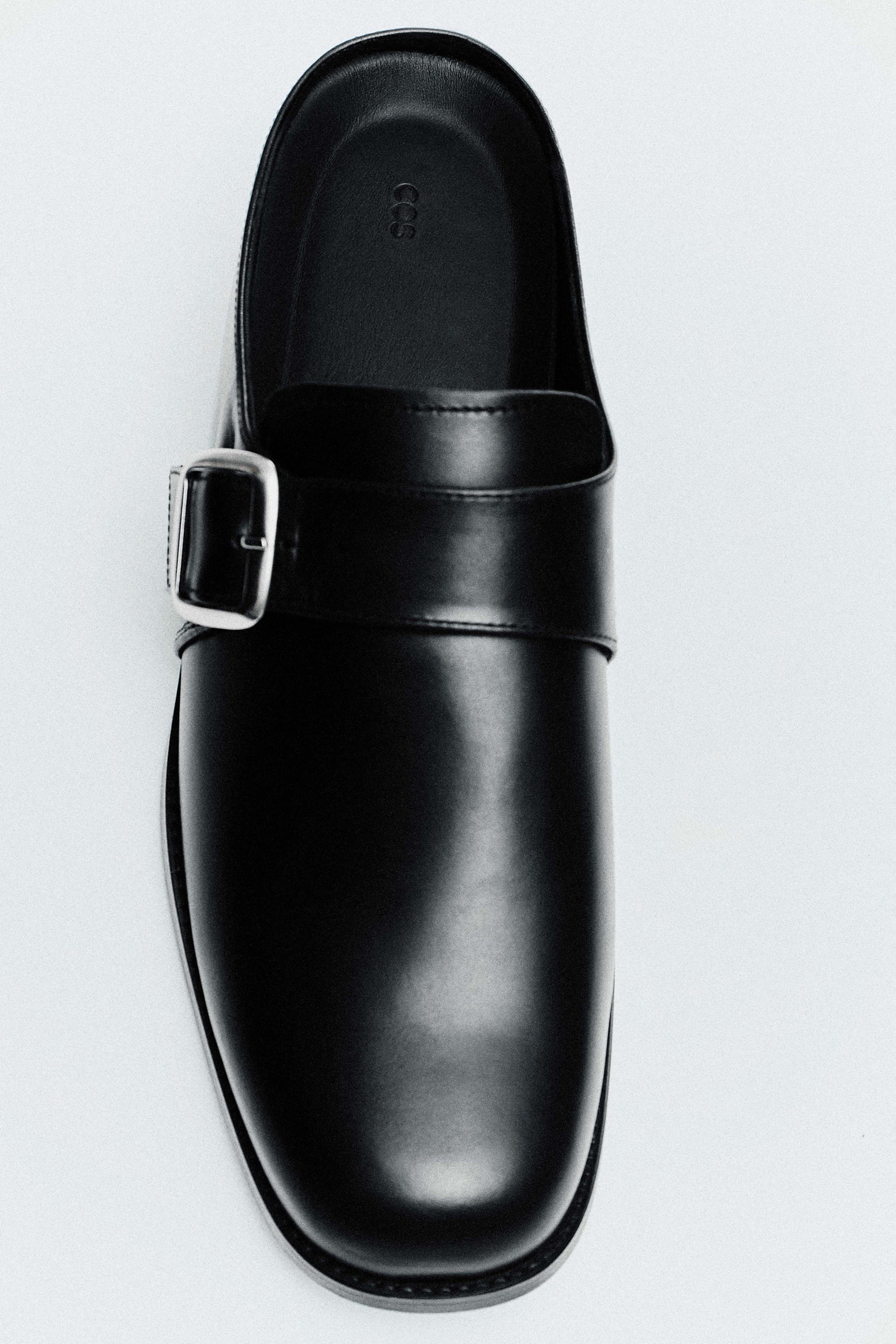 Größeres Bild ansehen: Ein Derby-Schuh aus Leder mit eckiger Zehenpartie liegt in der Draufsicht und zeigt ein backless Slip-on-Design aus glattem schwarzem Leder. Der Schuh verfügt über eine abgerundete eckige Zehenpartie, einen breiten Riemen über dem Spann mit polierter Schnalle in Silberoptik und ein geprägtes Logo auf dem schwarzen Fußbett.