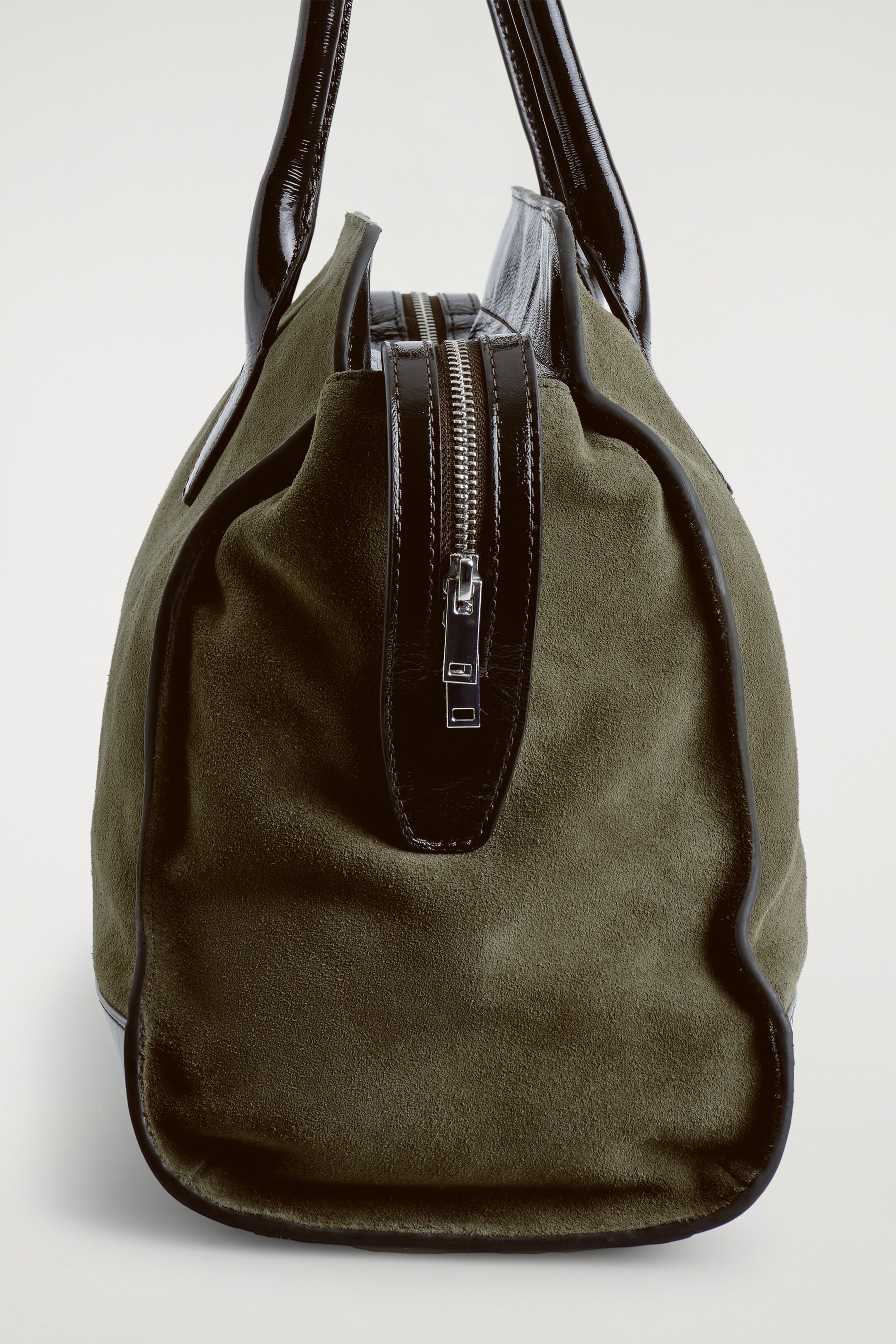 View larger image: A khaki suede tote bag is being viewed from a low angle, showing its rounded bottom structure. It is featuring shiny black patent leather handles and a central silver-toned two-way zipper closure, which is framed by black leather accents.