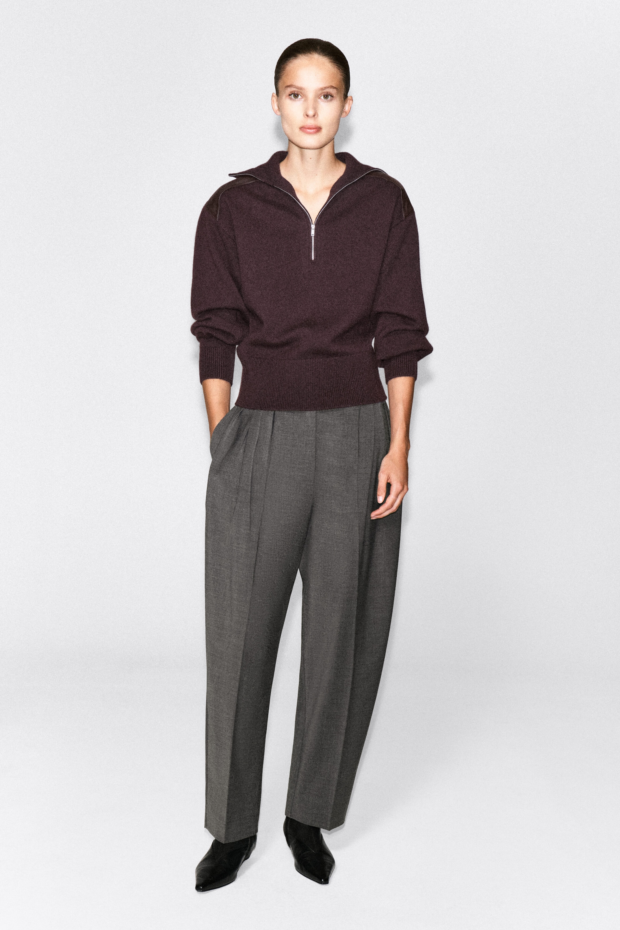 A dark burgundy merino wool zip-up sweater featuring a half-zip collar, ribbed trims, and subtle shoulder patches is worn by the model. The sweater is paired with gray pleated wide-leg trousers and black boots. A dark burgundy merino wool zip-up sweater featuring a half-zip collar, ribbed trims, and subtle shoulder patches is worn by the model. The sweater is paired with gray pleated wide-leg trousers and black boots.