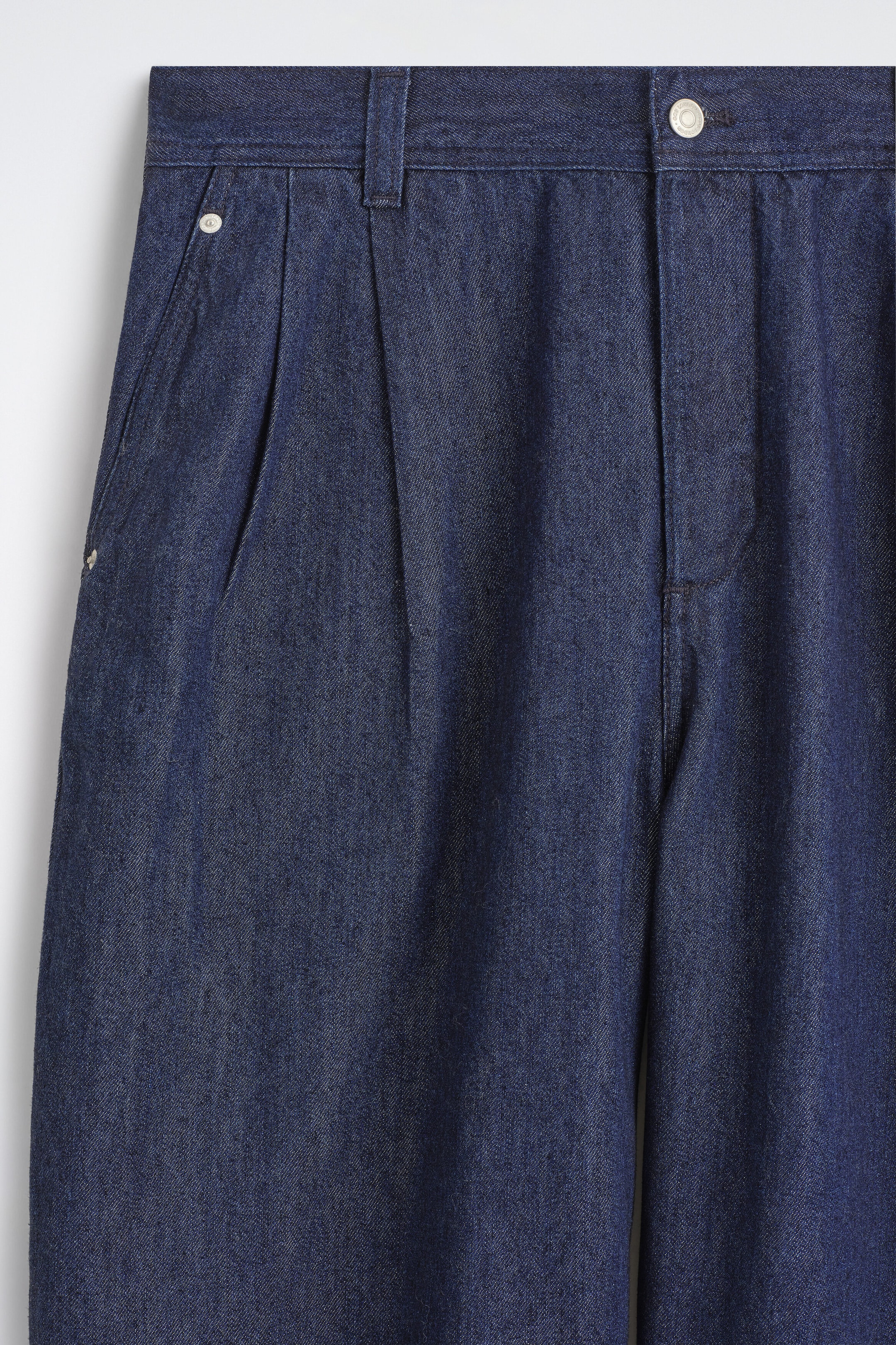Ce gros plan de face du pantalon barrel-leg en denim plissé coupe décontractée met en valeur un délavage bleu foncé, avec une ceinture à passants et une fermeture par bouton argenté. Le modèle présente des plis sur le devant ainsi qu’une poche latérale plaquée avec rivet métallique. Ce gros plan de face du pantalon barrel-leg en denim plissé coupe décontractée met en valeur un délavage bleu foncé, avec une ceinture à passants et une fermeture par bouton argenté. Le modèle présente des plis sur le devant ainsi qu’une poche latérale plaquée avec rivet métallique.