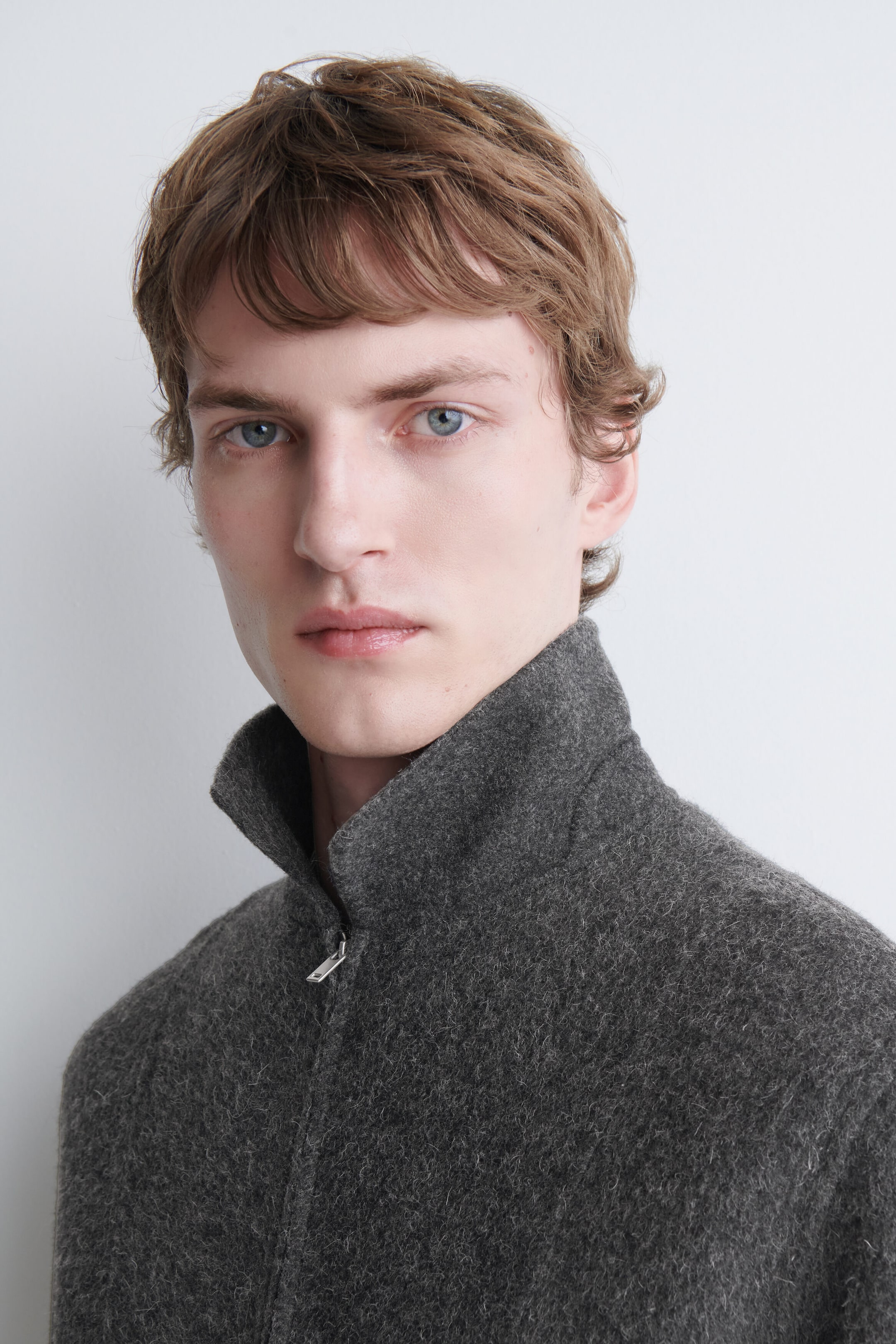 The model is wearing a dark gray, textured wool-like jacket with a high stand-up collar and a partial silver zipper.