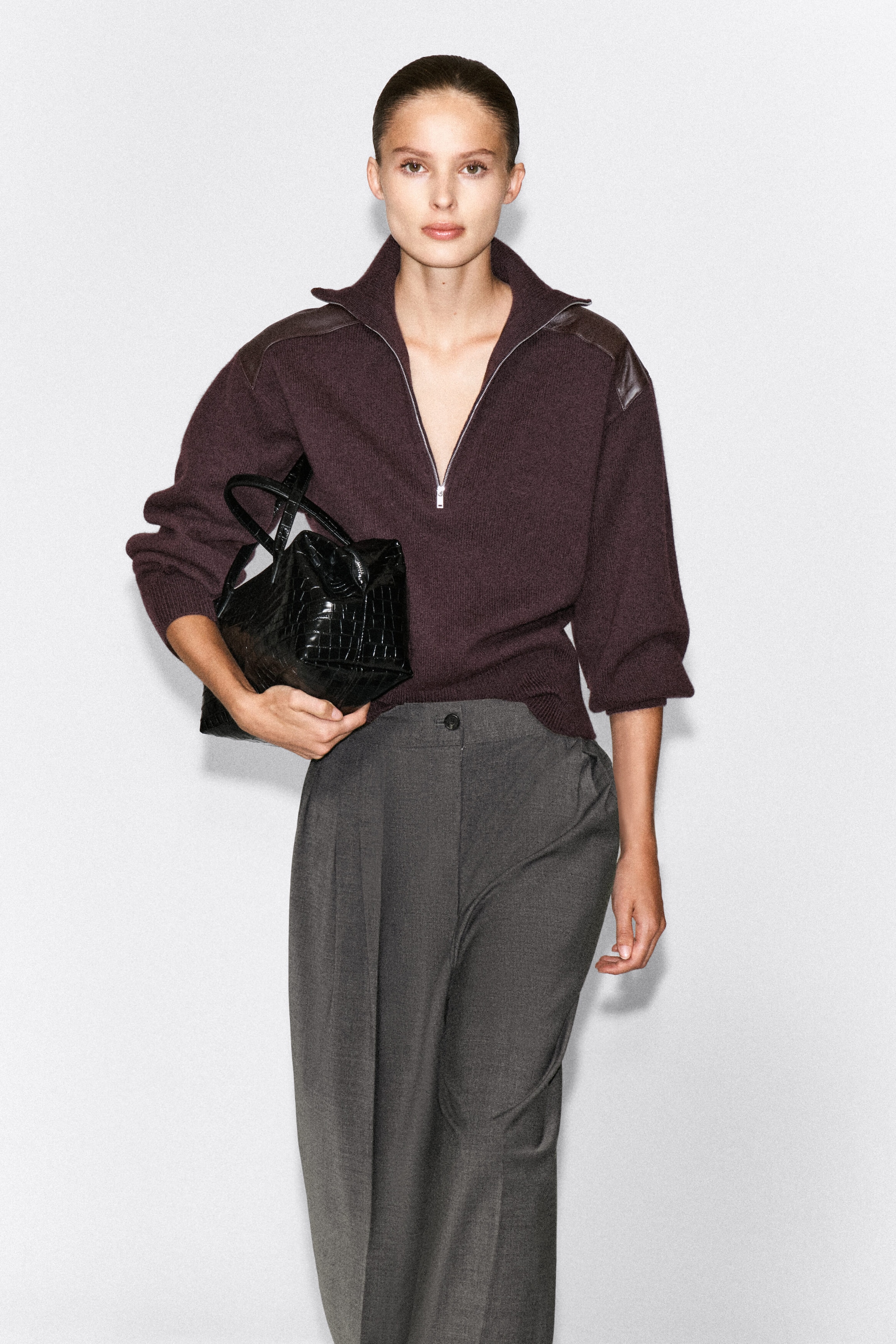 The dark plum leather-patch merino wool zip-up sweater is worn by the model and features a half-zip collar and matching leather shoulder panels. It is styled with grey wide-leg trousers, and the model is carrying a black crocodile-effect handbag under her arm. The dark plum leather-patch merino wool zip-up sweater is worn by the model and features a half-zip collar and matching leather shoulder panels. It is styled with grey wide-leg trousers, and the model is carrying a black crocodile-effect handbag under her arm.