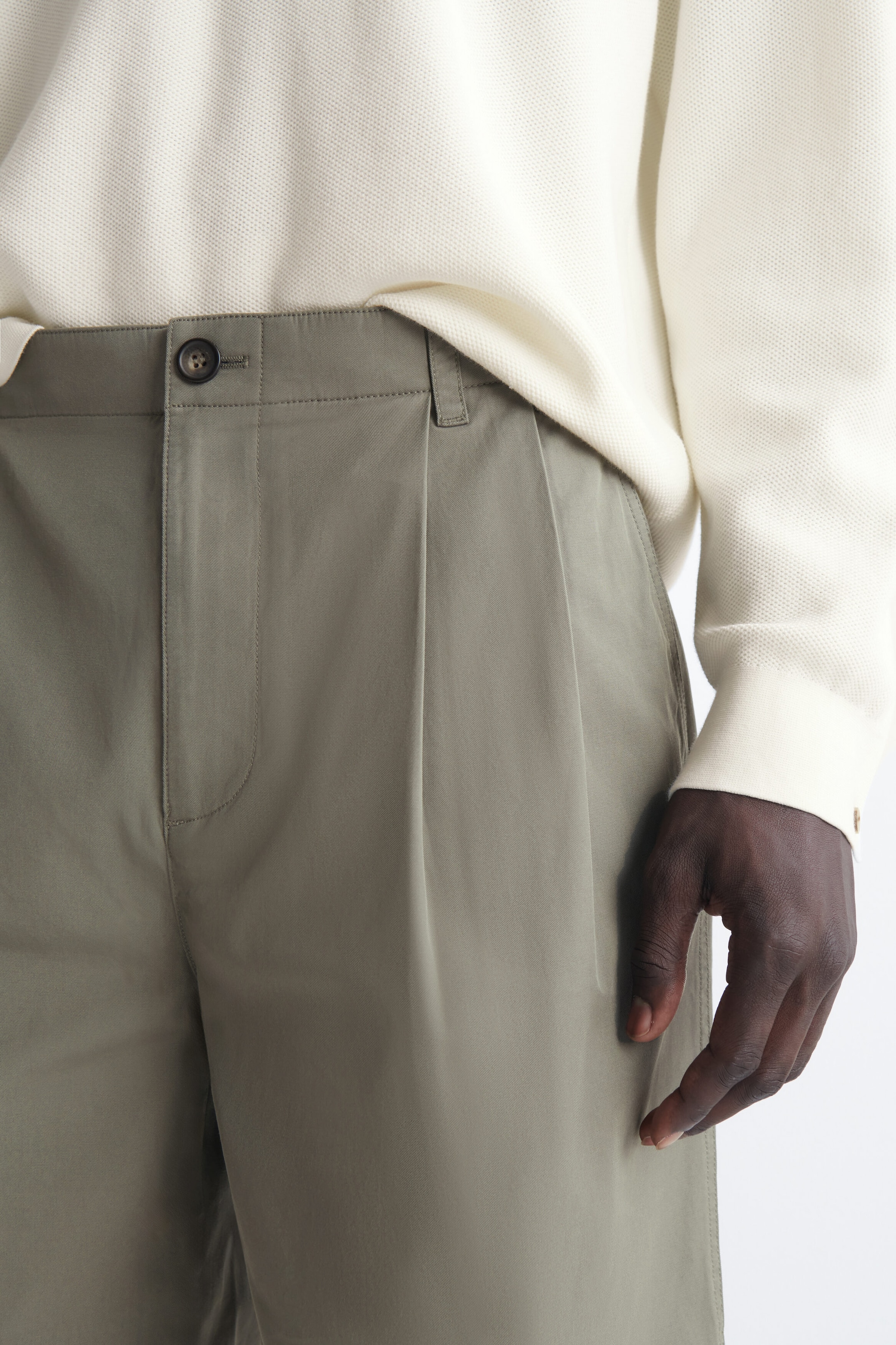 Maßgeschneiderte Falten-Shorts aus Baumwolle in Salbeigrün sind in der oberen Frontansicht zu sehen und verfügen über einen strukturierten Bund mit Einzelknopfverschluss, Gürtelschlaufen und eine ausgeprägte vordere Falte. Ein cremefarbenes Langarm-Top mit strukturierter Oberfläche ist teilweise in den Bund gesteckt.