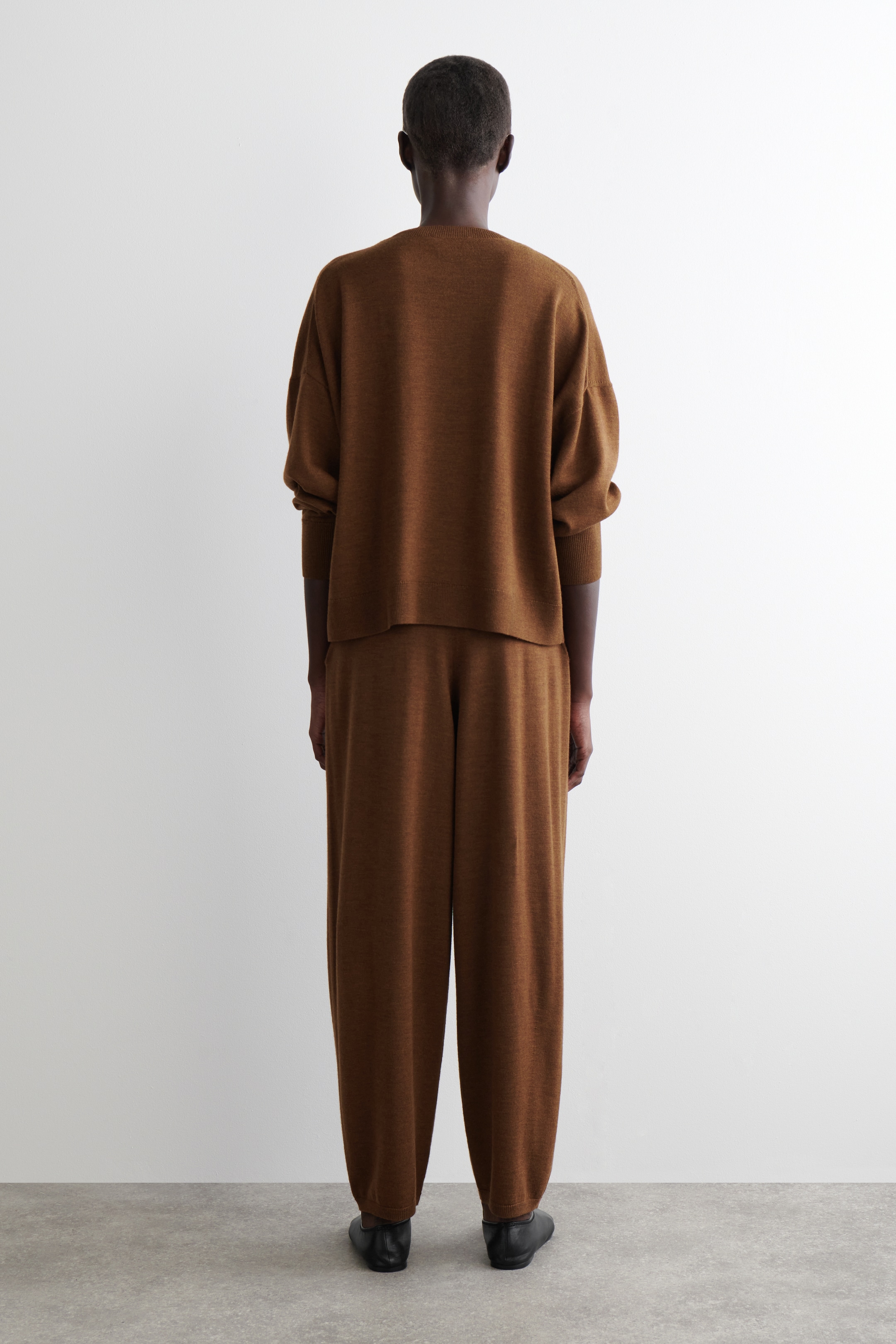 Visualizza immagine più grande: Un set coordinato marrone lavorato a maglia viene mostrato da dietro, caratterizzato da un top ampio e pantaloni a gamba larga coordinati. Il top presenta una silhouette rilassata con maniche lunghe che terminano in polsini a coste, mentre i pantaloni mostrano una caduta confortevole, con gli orli che poggiano su scarpe basse nere.