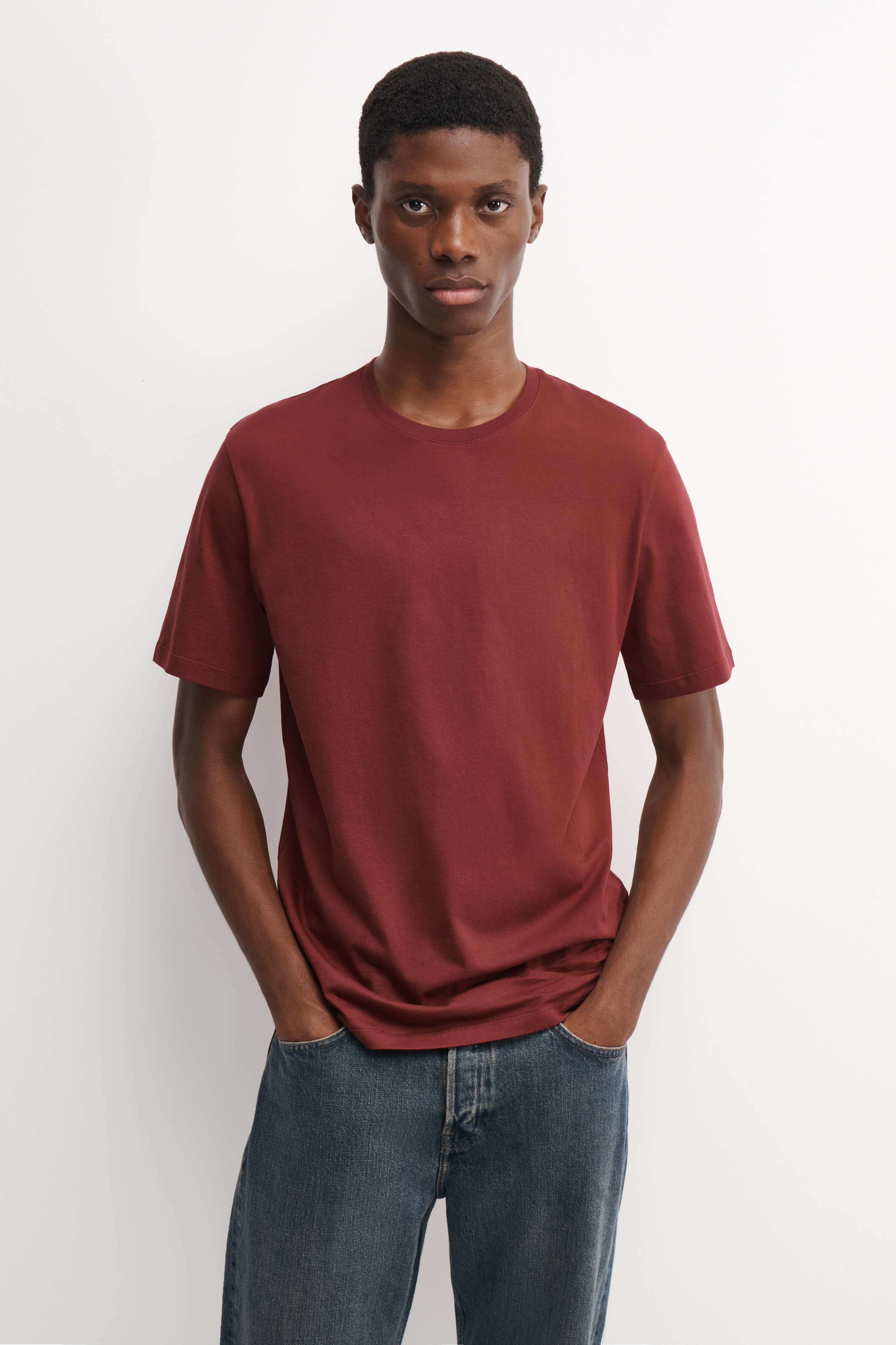 Three cotton crew-neck T-shirts are overlapping in a diagonal row, facing front and displaying their classic fit and structure. The short-sleeve T-shirts feature ribbed crew necklines and straight hems, shown in sage green, dark purple and brick red.