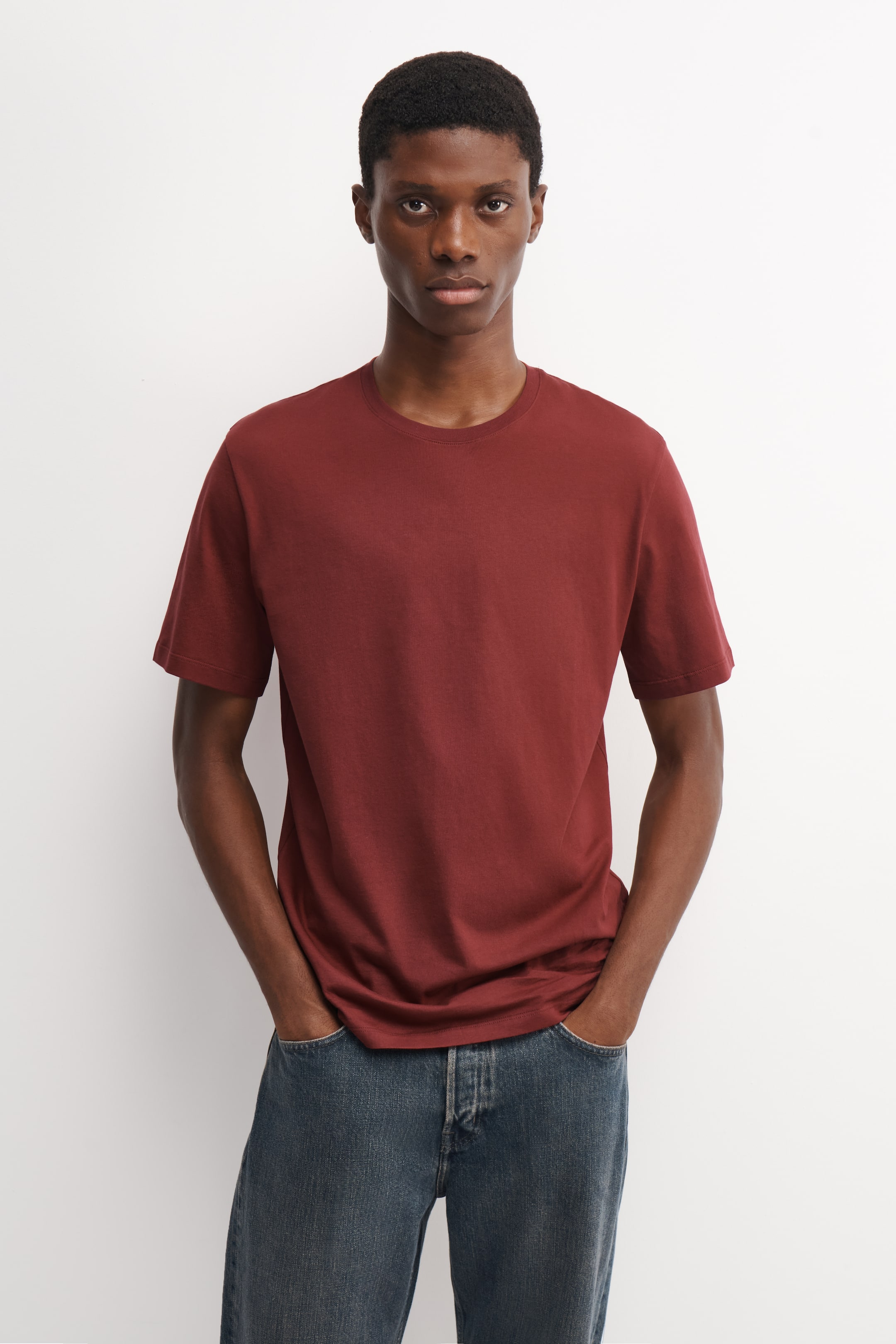 Ver imagen más grande: La modelo lleva puesta una camiseta de cuello redondo de algodón en rojo oscuro, posando con las manos en los bolsillos de unos vaqueros azules. La camiseta de manga corta presenta un corte clásico con escote acanalado.