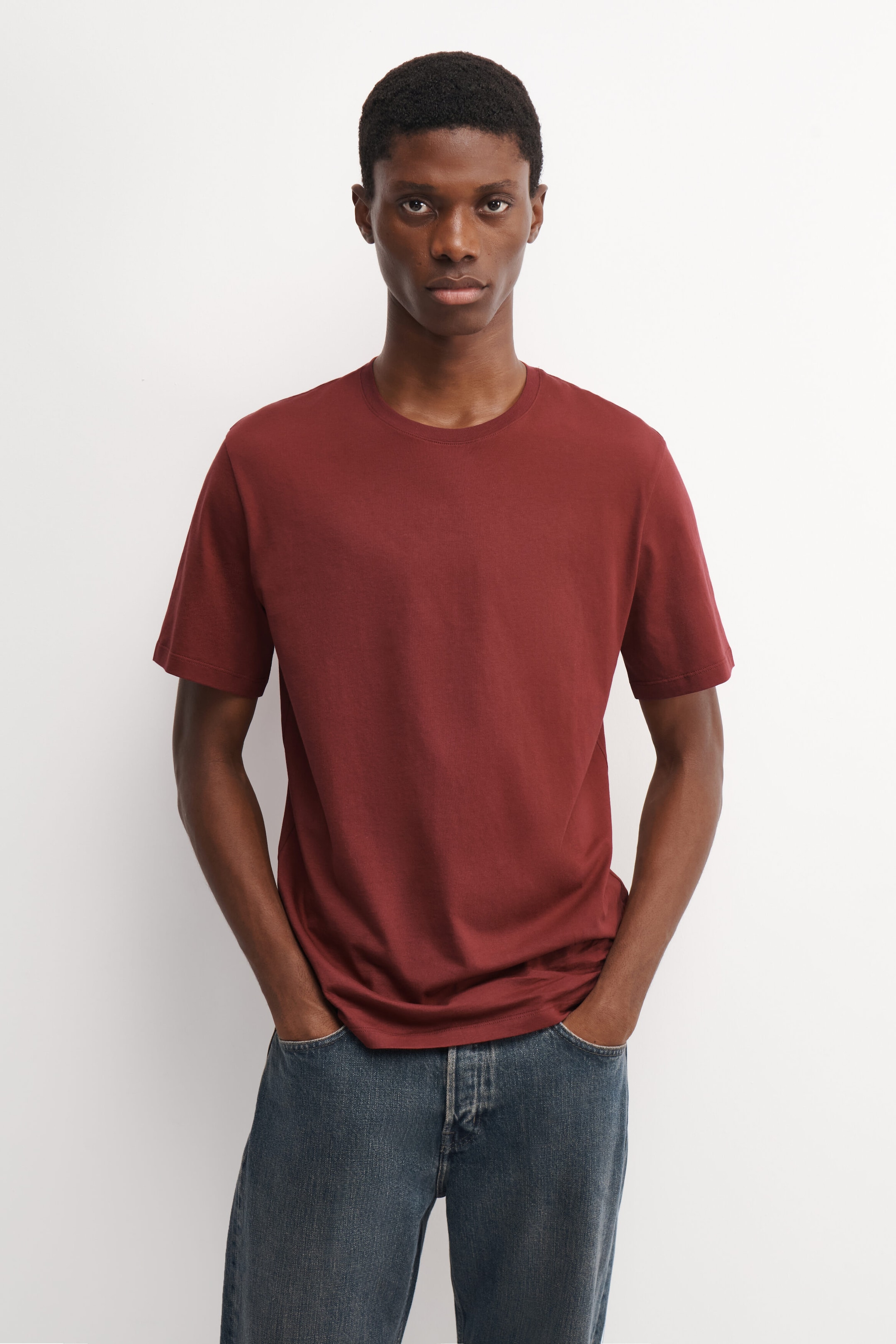 A dark red cotton crew-neck T-shirt is being worn by the model, who is posing with hands in the pockets of blue jeans. The short-sleeved T-shirt features a classic fit with a ribbed neckline. A dark red cotton crew-neck T-shirt is being worn by the model, who is posing with hands in the pockets of blue jeans. The short-sleeved T-shirt features a classic fit with a ribbed neckline.