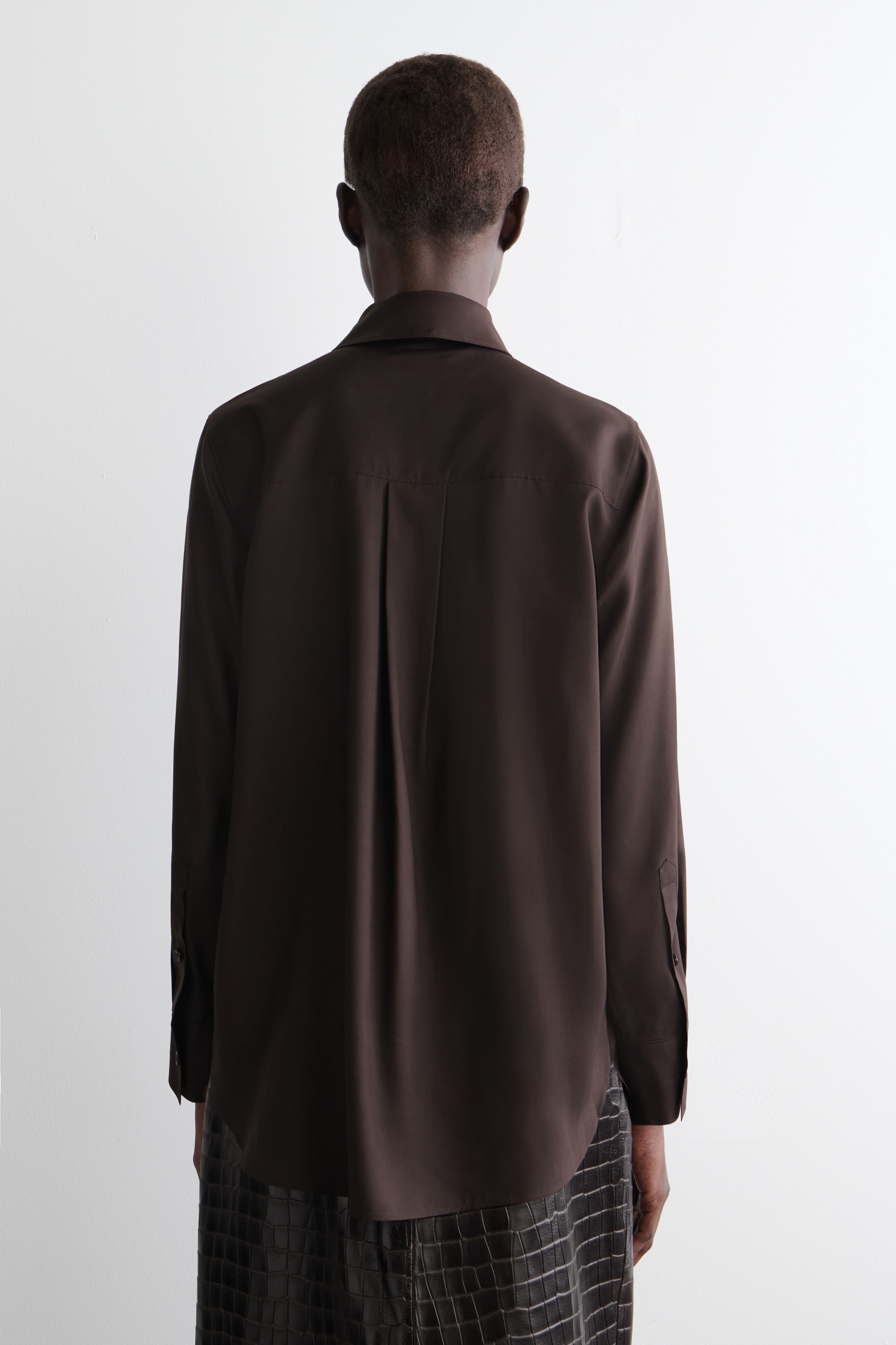 Ver imagen más grande: Una camisa marrón oscuro se presenta desde atrás, con un cuello estándar, un pliegue central que se extiende desde el canesú trasero y mangas largas con puños abotonados, mientras que su dobladillo curvado es visible sobre una falda estampada.