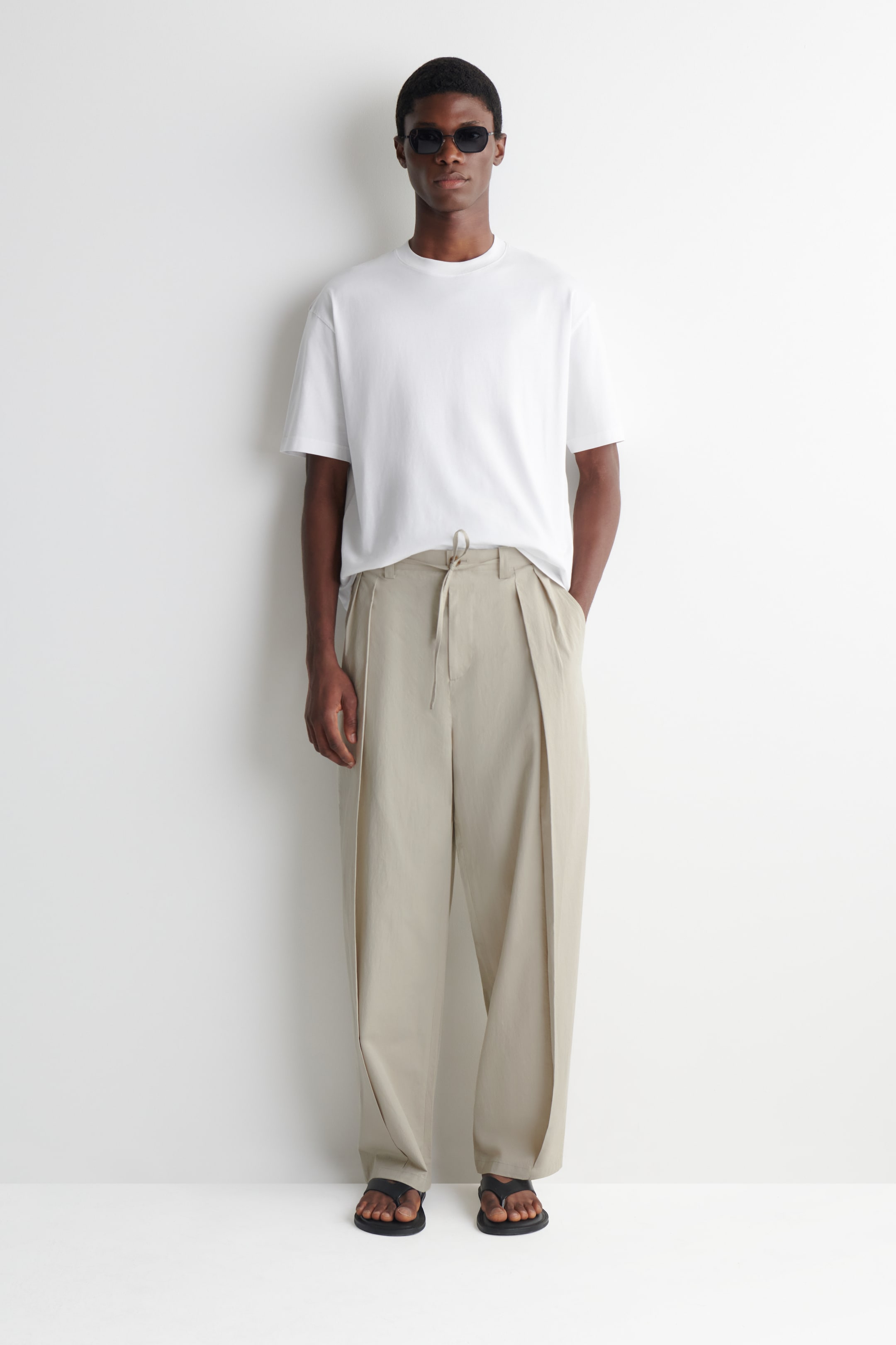Ver imagen más grande: Pantalones de pierna ancha en color beige claro, con cintura ajustable mediante cordón y prominentes pliegues dobles que recorren cada pierna. Caen rectos desde la cintura, con el cordón visible sobre una camiseta blanca metida por dentro, y sus amplias perneras caen ligeramente sobre unas sandalias oscuras.