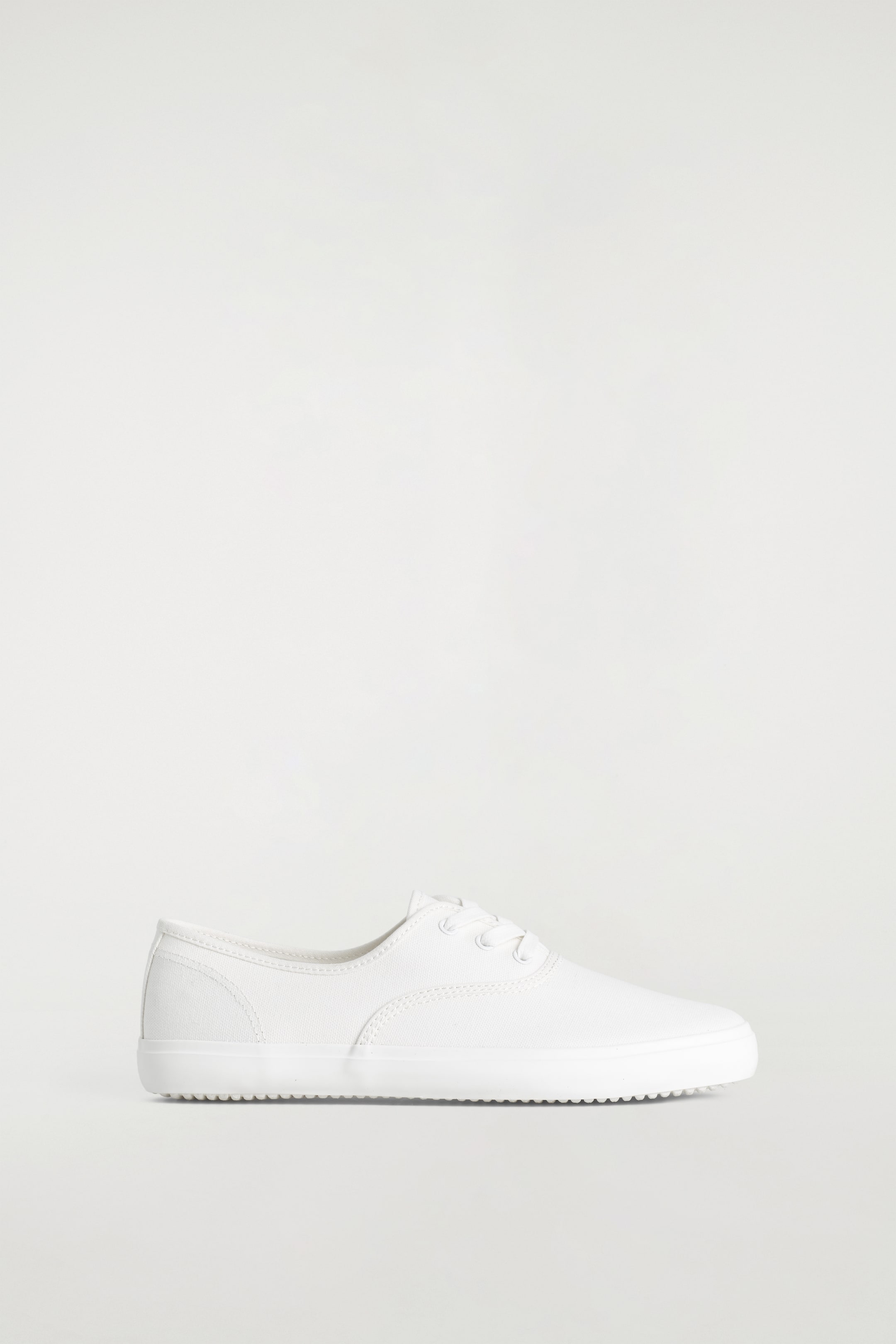 Agrandir l'image: Une chaussure en toile blanche à tige basse est orientée vers la droite, avec des lacets blancs passant à travers des œillets métalliques et des coutures visibles sur sa partie supérieure. Sa semelle en caoutchouc blanc présente un dessous texturé.