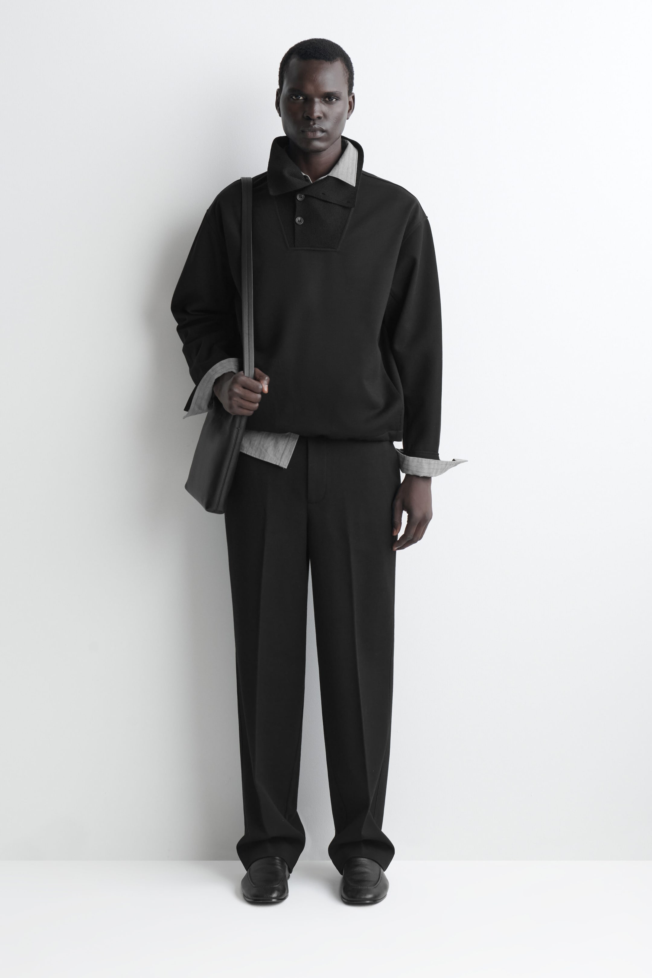 A model is standing facing forward, wearing a black funnel-neck pullover with a buttoned placket layered over a grey collared shirt, with the cuffs turned back. The look is completed with black wide-leg trousers featuring a sharp front crease, black leather slip-on shoes, and a black leather shoulder bag.
