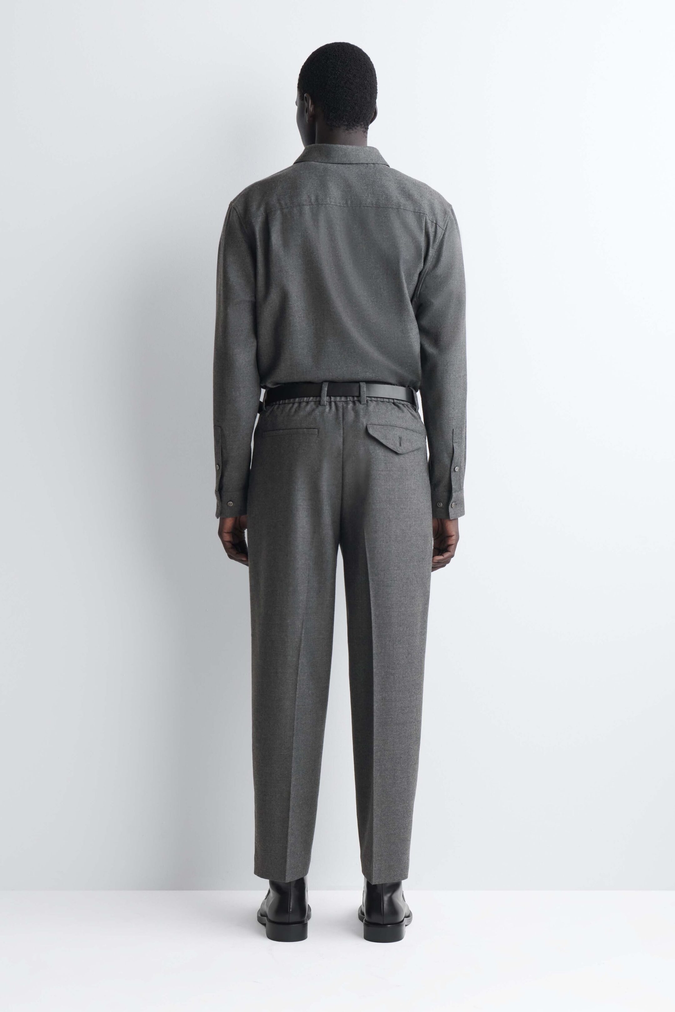 Das passende graue Langarmhemd und die graue Hose sind von hinten zu sehen, wobei das Hemd in die Hose gesteckt und mit einem schwarzen Gürtel fixiert ist. Die Hose mit geradem Bein zeigt mittig gebügelte Bügelfalten entlang der Beine, eine Paspeltasche links und eine Pattentasche mit Knopf rechts. Das passende graue Langarmhemd und die graue Hose sind von hinten zu sehen, wobei das Hemd in die Hose gesteckt und mit einem schwarzen Gürtel fixiert ist. Die Hose mit geradem Bein zeigt mittig gebügelte Bügelfalten entlang der Beine, eine Paspeltasche links und eine Pattentasche mit Knopf rechts.
