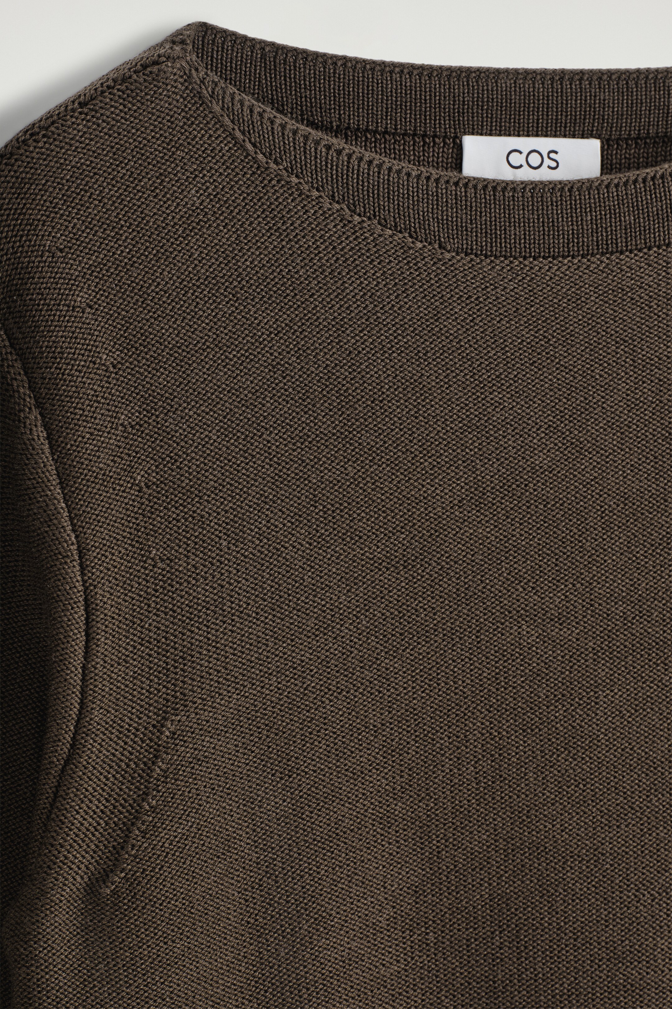 Agrandir l'image: Un pull en tricot marron foncé est présenté de face, avec un col rond côtelé et une texture de tricot fine et dense, avec une étiquette blanche affichant 'COS' visible à l'arrière du cou.