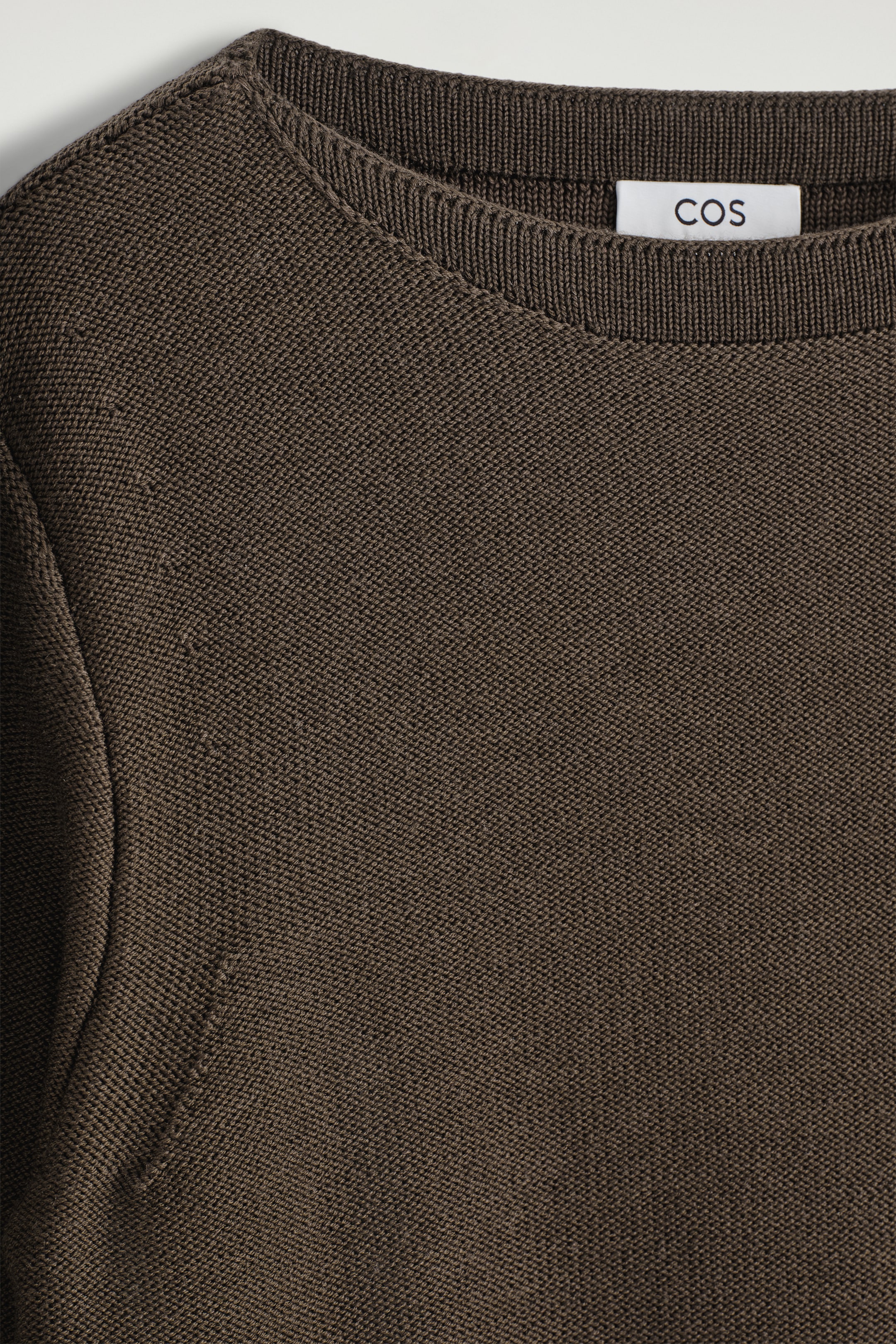 Grösseres Bild ansehen: Ein dunkelbrauner Strickpullover wird von vorne gezeigt, mit einem gerippten Rundhalsausschnitt und einer feinen, dichten Strickstruktur, mit einem weißen Etikett, das 'COS' zeigt, sichtbar am hinteren Teil des Halses.