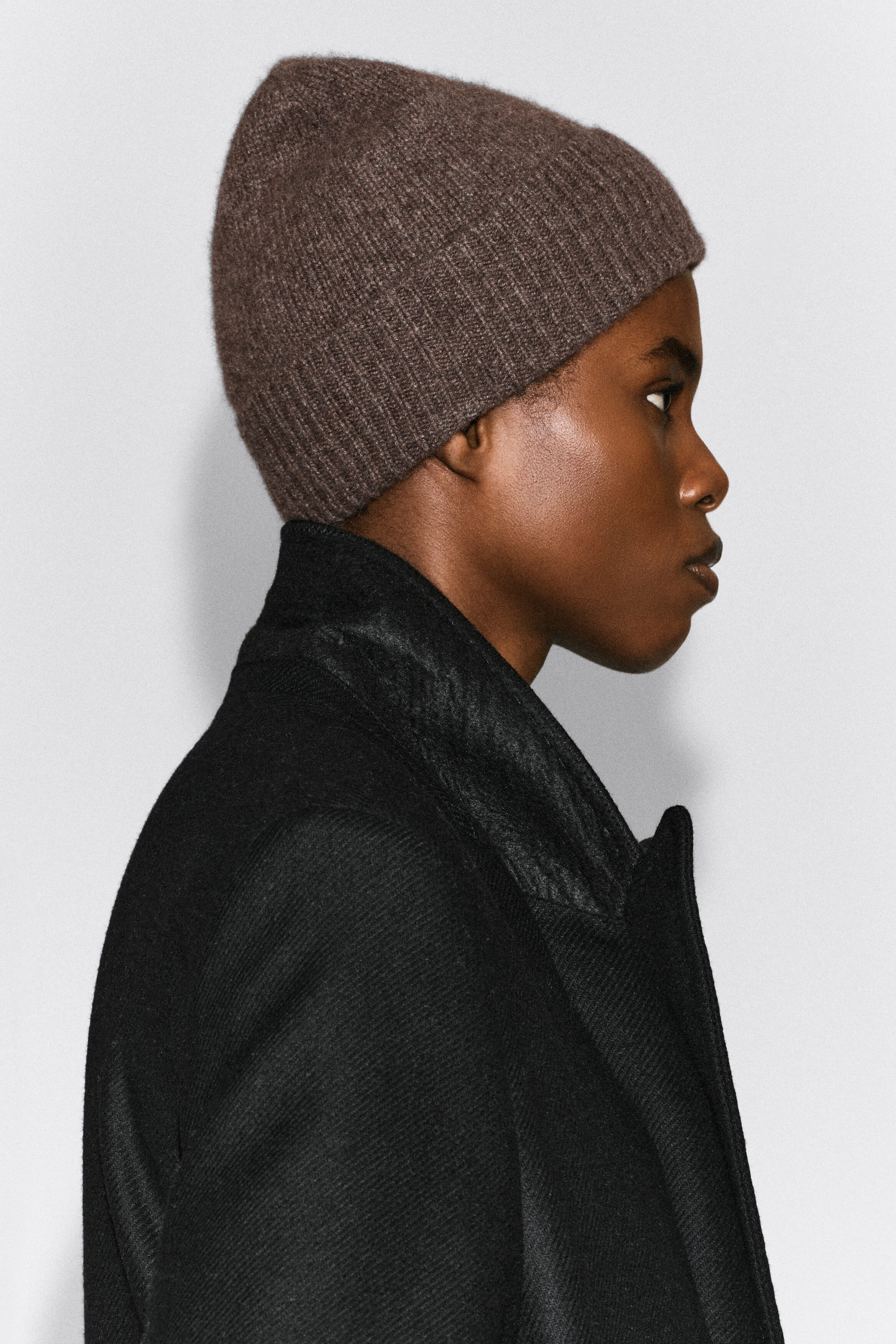 Un bonnet en cachemire brun avec revers côtelé est porté par le mannequin, montré de profil. Le bonnet est stylisé avec un manteau texturé noir.