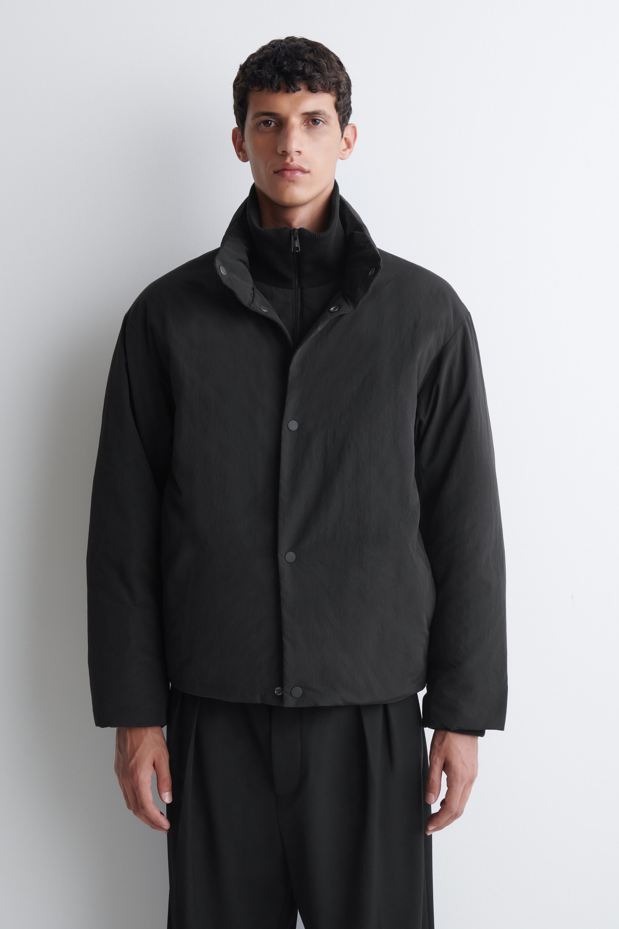 A padded funnel-neck down jacket in black is worn by the model, featuring a snap-button front closure and a high collar. The jacket is layered over a black ribbed zip-up sweater and black trousers. A padded funnel-neck down jacket in black is worn by the model, featuring a snap-button front closure and a high collar. The jacket is layered over a black ribbed zip-up sweater and black trousers.