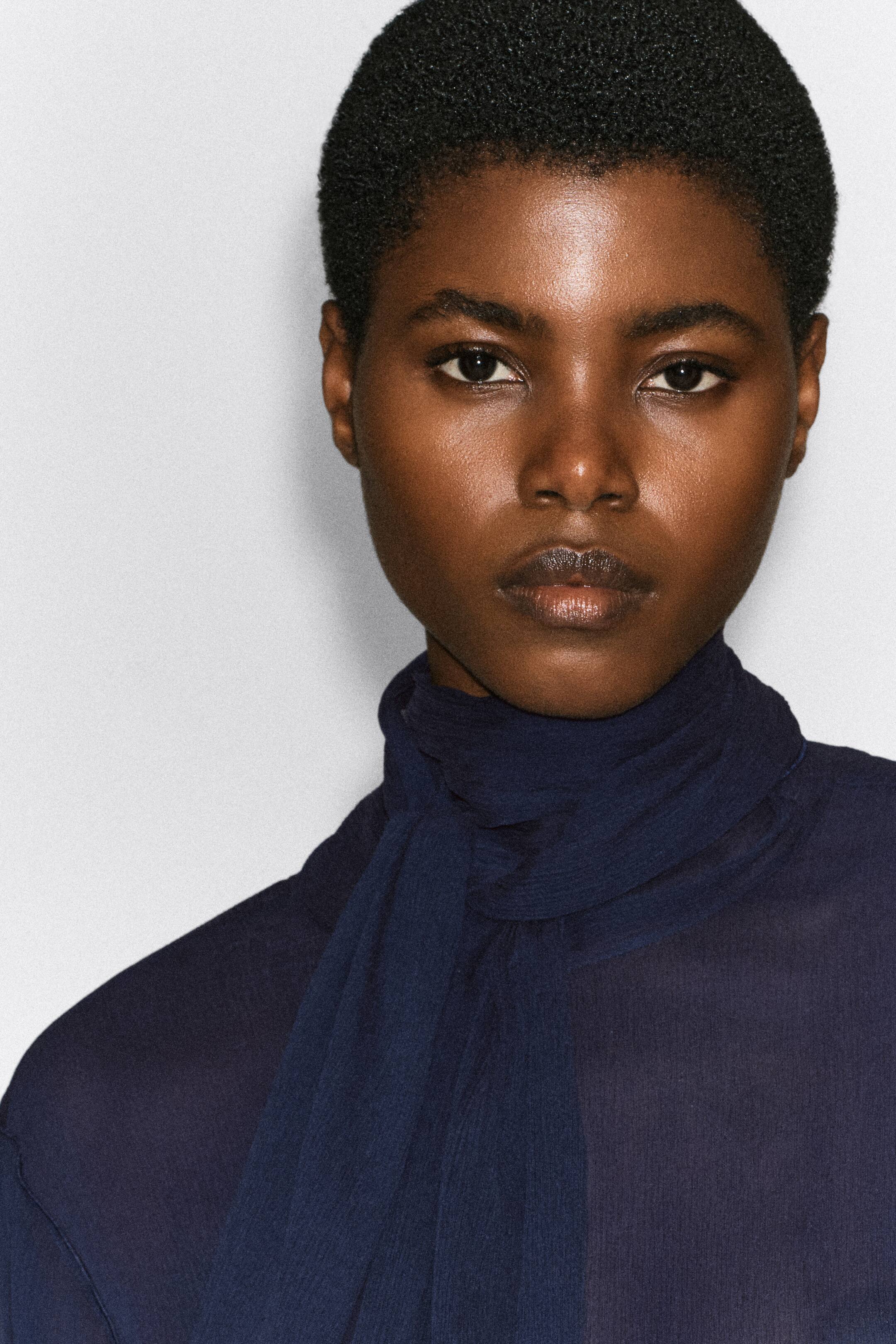 View larger image: The model is wearing a dark blue, semi-sheer crepe top, featuring a matching scarf wrapped around the neck and draped down the front.