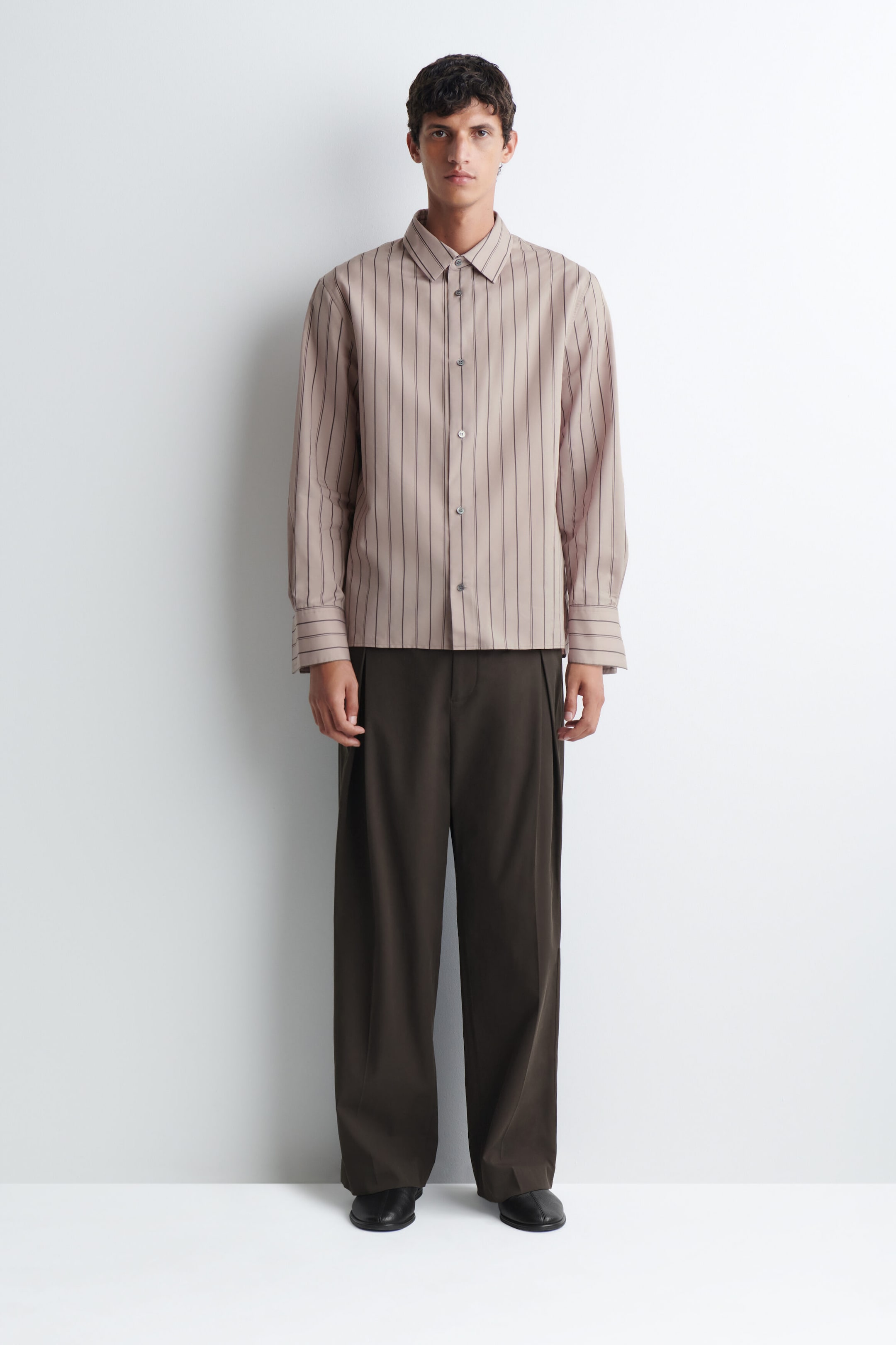 A striped structured cotton-silk shirt in beige with dark vertical stripes is being worn by the model. The shirt features a pointed collar, a button-down front, and long sleeves with wide cuffs, paired with dark brown wide-leg trousers. A striped structured cotton-silk shirt in beige with dark vertical stripes is being worn by the model. The shirt features a pointed collar, a button-down front, and long sleeves with wide cuffs, paired with dark brown wide-leg trousers.