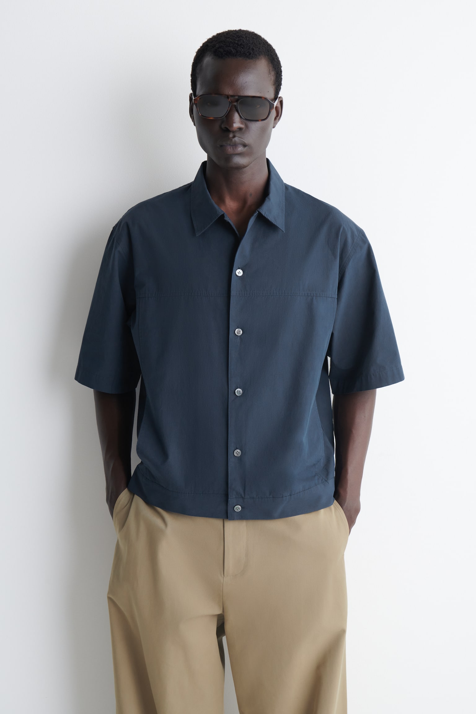 Una camicia da uomo blu scuro è rivolta frontalmente, caratterizzata da un colletto e maniche corte. È tagliata in una forma squadrata, con chiusura a bottoni bianchi e cuciture a vista che formano un pannello orizzontale sul petto e pannelli verticali lungo i lati. - 1