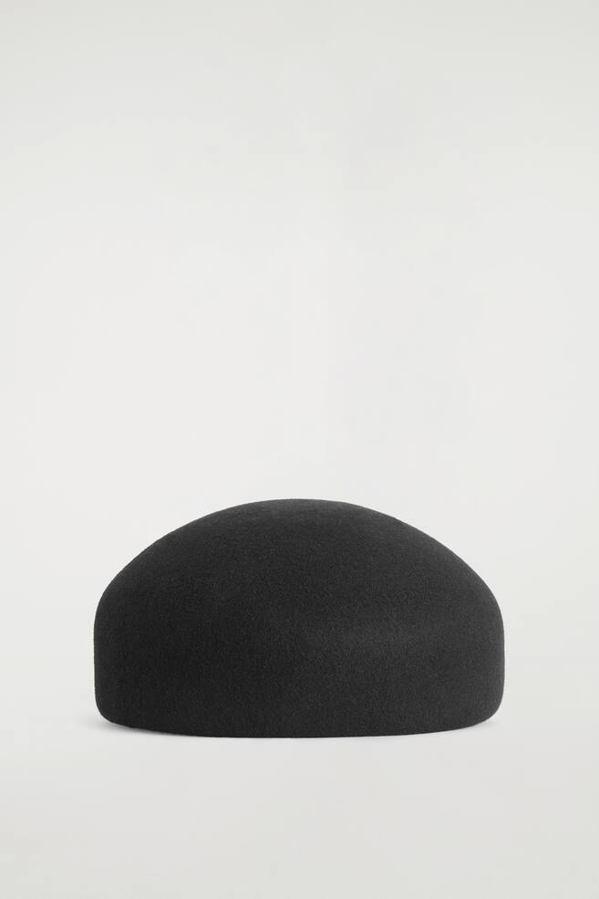 A black sculpted wool beret is resting on a flat surface, displaying its structured, dome-like silhouette and soft, felted texture from a front-facing view. - 1
