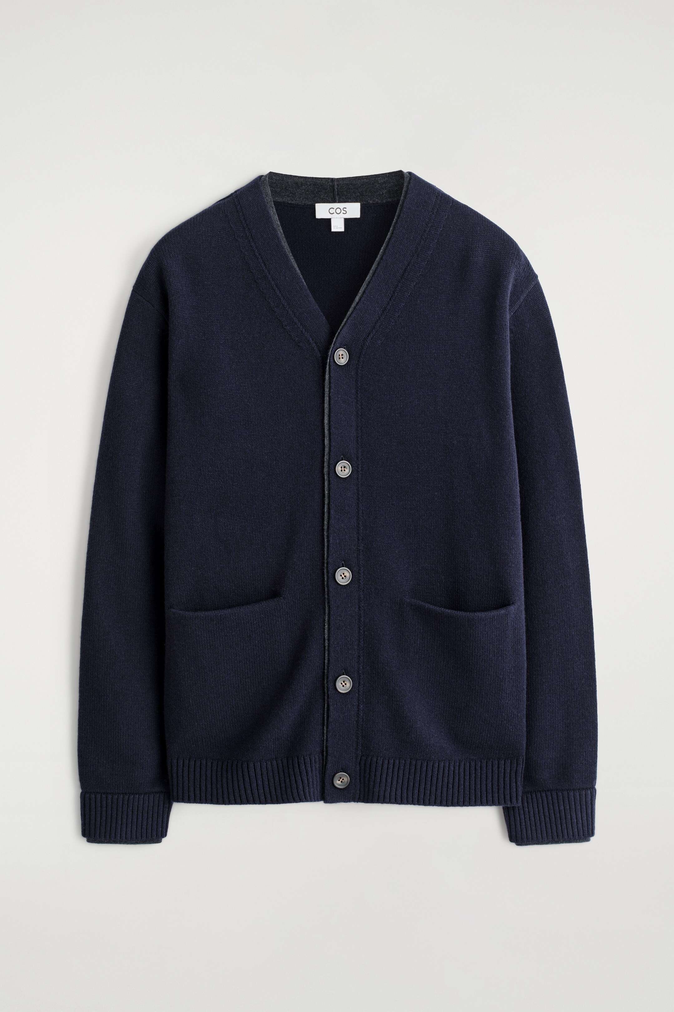 Grösseres Bild ansehen: Ein marineblauer Cardigan wird von vorne präsentiert, mit einem V-Ausschnitt mit kontrastierendem grauem Besatz, einem Knopfverschluss mit fünf dunklen, runden Knöpfen, zwei aufgesetzten Taschen auf der unteren Vorderseite und gerippten Bündchen und Saum. Ein dunkelgraues Rundhals-Shirt ist darunter geschichtet.