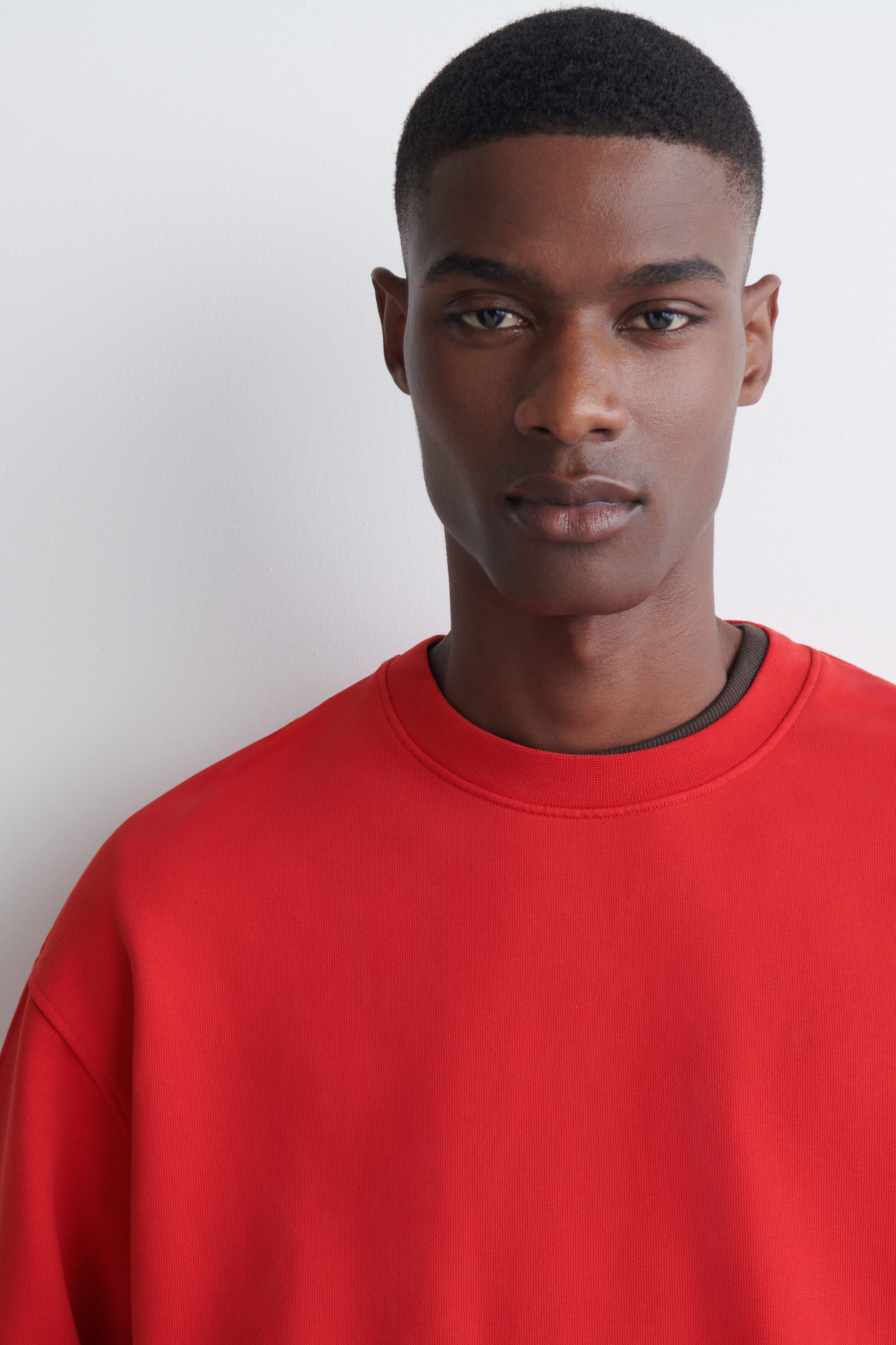 The model is wearing a red crew-neck sweatshirt with a ribbed neckline, layered over a dark collar. The model is wearing a red crew-neck sweatshirt with a ribbed neckline, layered over a dark collar.
