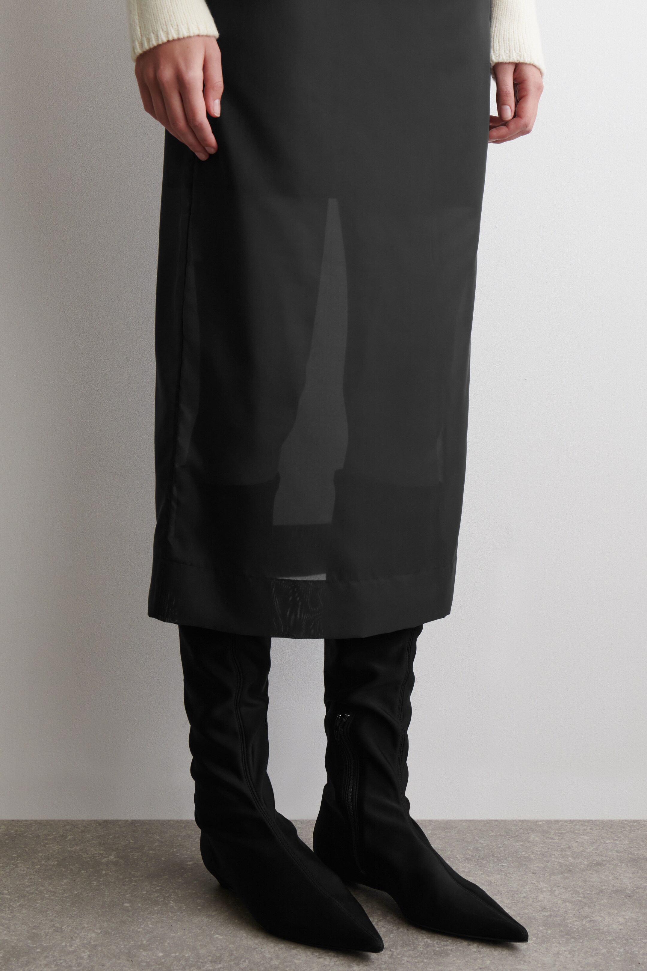 View larger image: A black column skirt is being presented from the FRONT, featuring a sheer outer layer over an opaque internal layer. The opaque internal layer ends above the knee, revealing the sheer fabric extending to the skirt's midi-length hem.