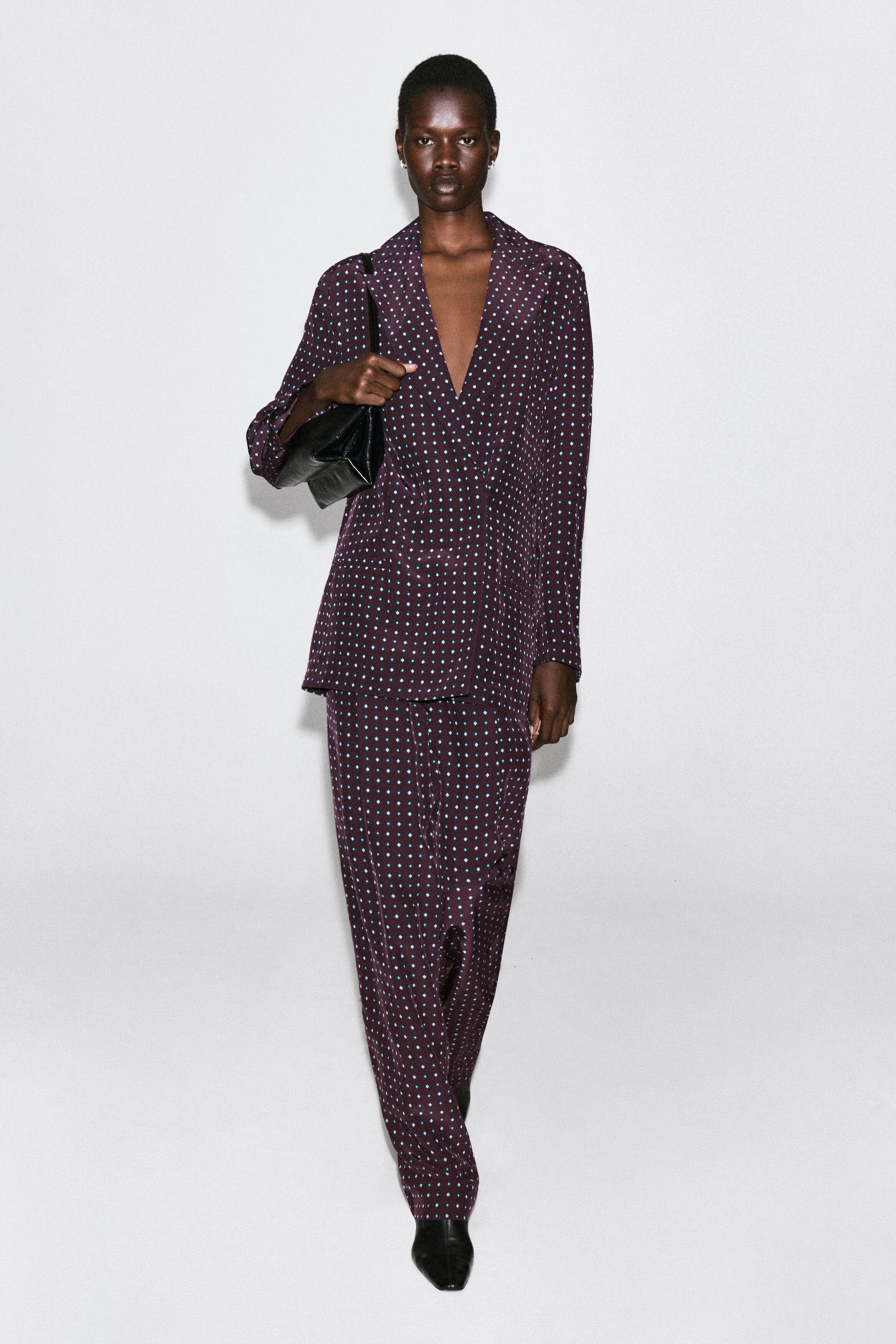 A front view showcases a deep purple, diamond-print double-breasted blazer featuring notch lapels and a relaxed, fluid drape. The blazer is styled with matching trousers and a black shoulder bag held under the arm, which gently gathers the sleeve. A front view showcases a deep purple, diamond-print double-breasted blazer featuring notch lapels and a relaxed, fluid drape. The blazer is styled with matching trousers and a black shoulder bag held under the arm, which gently gathers the sleeve.