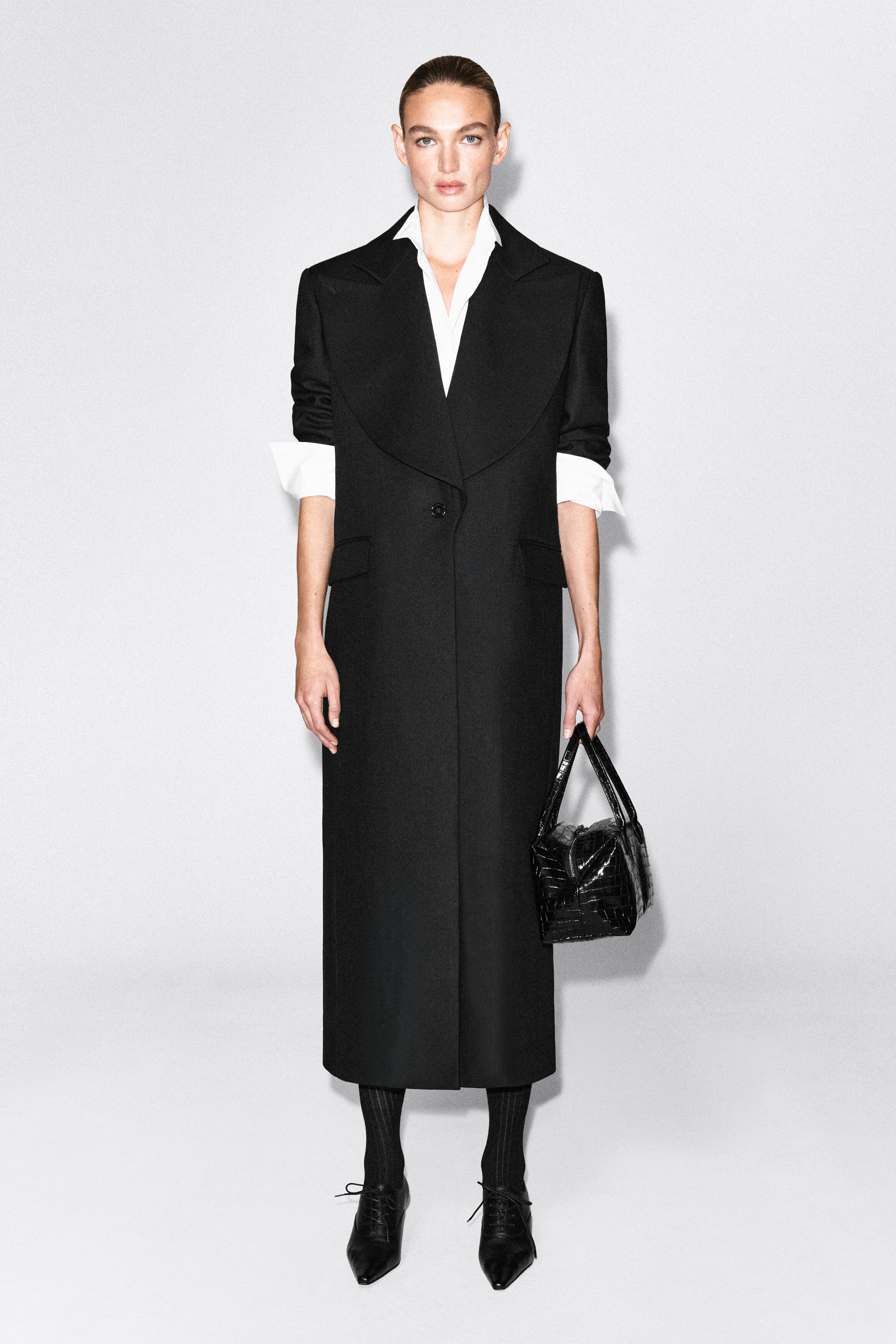 A long black single-breasted wool coat with wide draped lapels and a single-button fastening is worn by the model. The coat is layered over a white collared shirt with turned-back cuffs, and the model is carrying a black textured handbag.