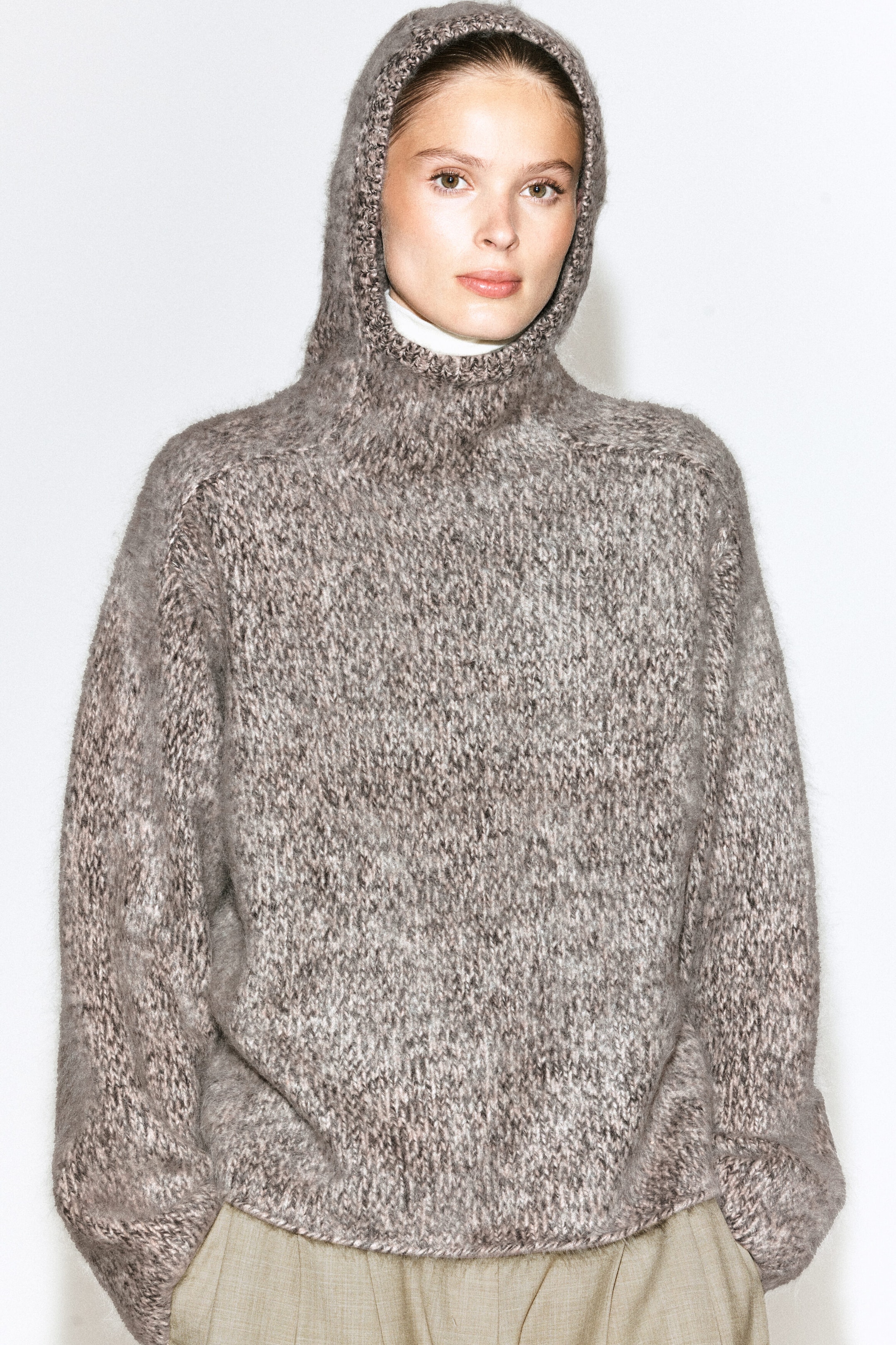 A chunky brushed-cashmere hoodie in a textured, melange brown knit is being worn by the model with the integrated hood pulled up over her head. The oversized, long-sleeved knitwear is layered over a white turtleneck top and styled with light beige trousers. A chunky brushed-cashmere hoodie in a textured, melange brown knit is being worn by the model with the integrated hood pulled up over her head. The oversized, long-sleeved knitwear is layered over a white turtleneck top and styled with light beige trousers.