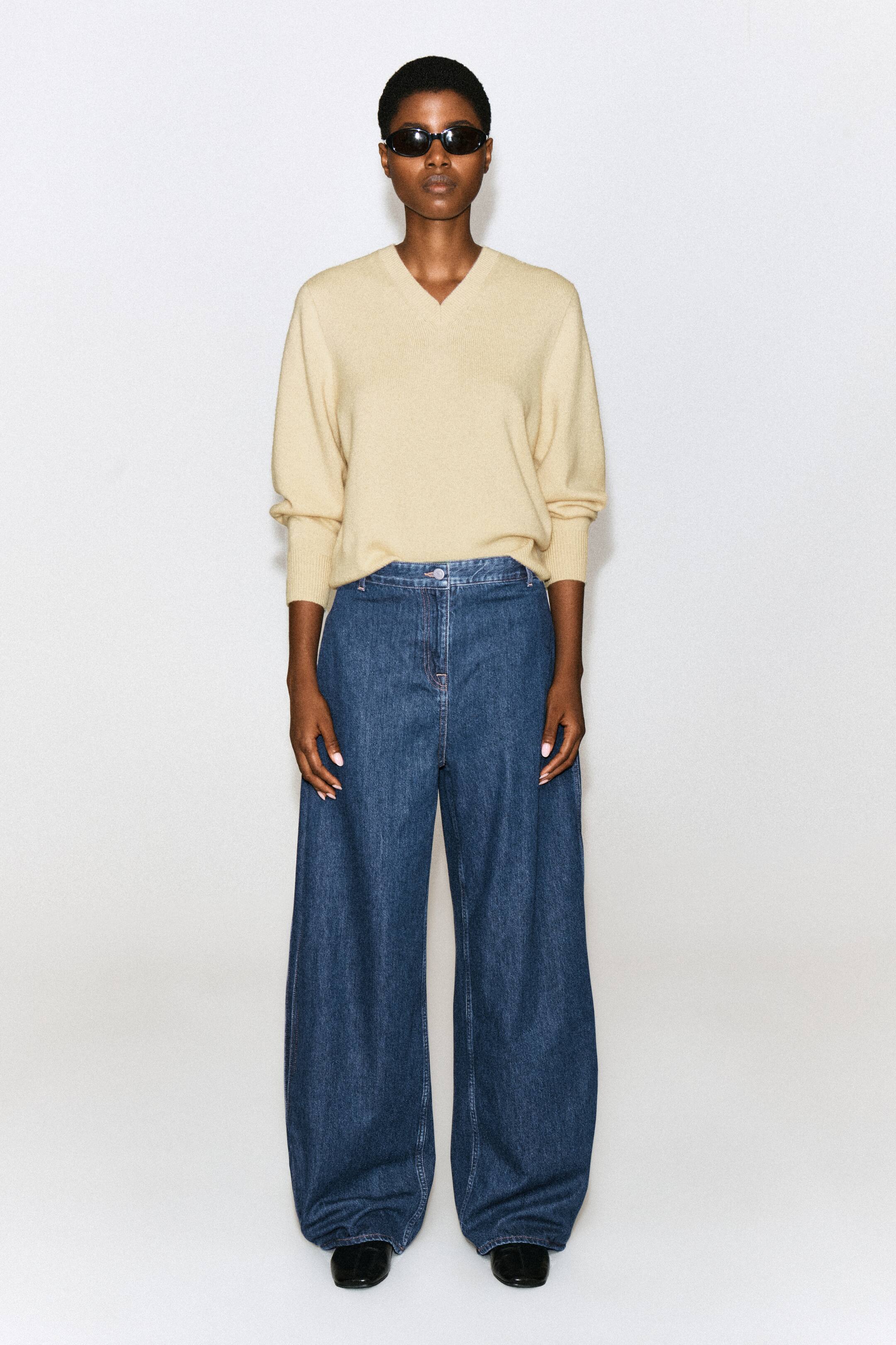 Agrandir l'image: Un pantalon barrel en denim à panneaux plissés, délavage bleu moyen, est porté par le mannequin, mettant en valeur une taille haute et une silhouette à jambe large et arrondie. Le pantalon est associé à un pull beige clair à col V rentré à la taille et à des chaussures noires.