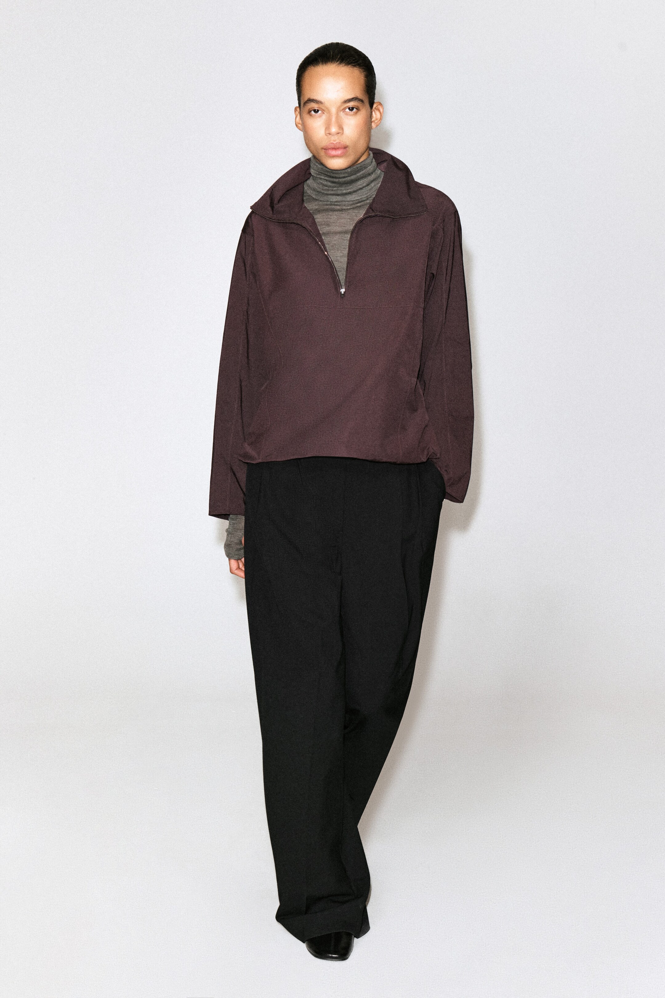 The dark plum half-zip pullover is shown from the front, featuring a relaxed fit, a silver-tone zipper, and a high collar. It is layered over a grey turtleneck top and paired with wide-leg black trousers. The dark plum half-zip pullover is shown from the front, featuring a relaxed fit, a silver-tone zipper, and a high collar. It is layered over a grey turtleneck top and paired with wide-leg black trousers.