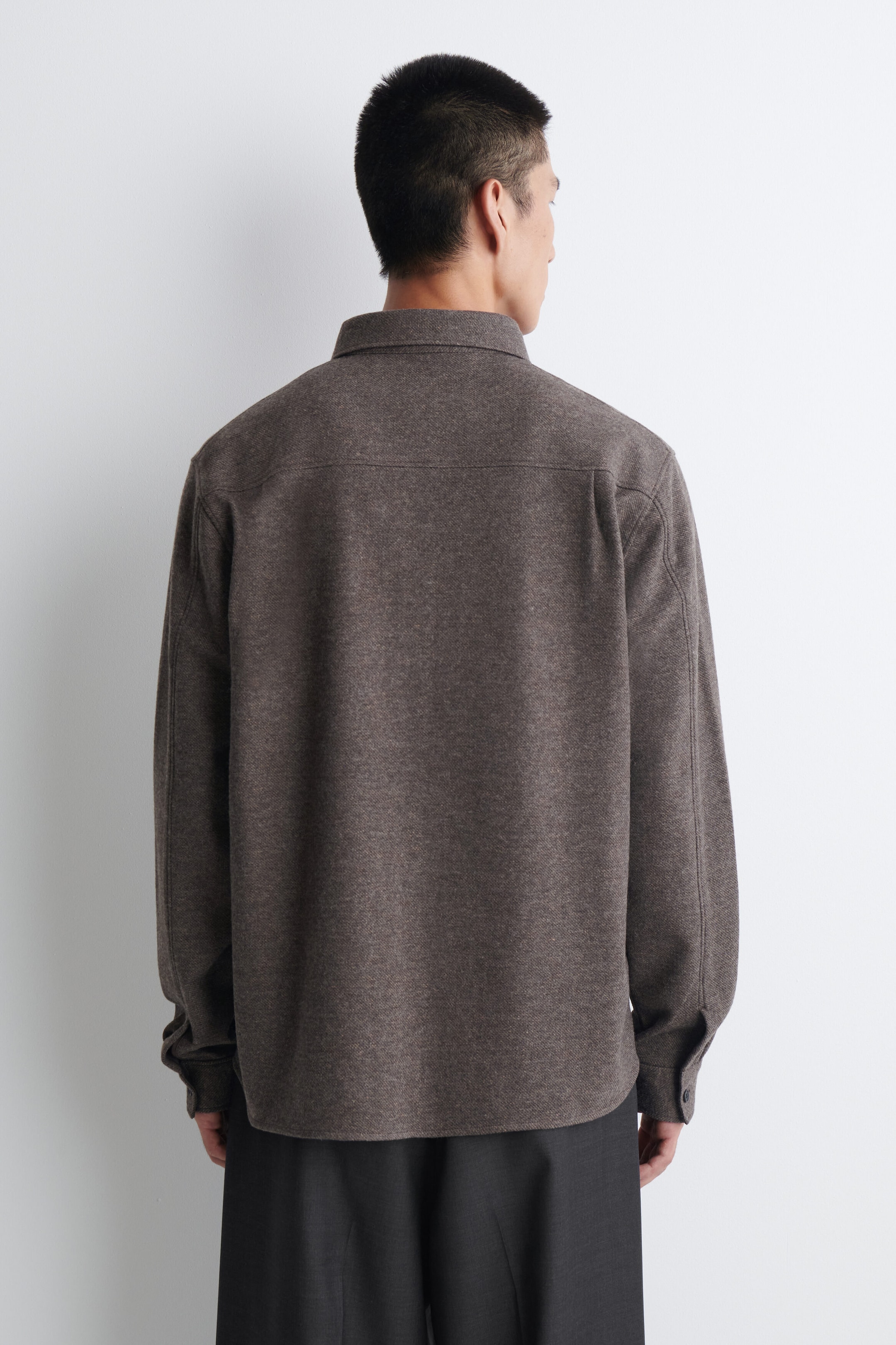 A dark brown textured overshirt is shown from the back, featuring a classic collar, a distinctive back yoke, and long sleeves with buttoned cuffs. A dark brown textured overshirt is shown from the back, featuring a classic collar, a distinctive back yoke, and long sleeves with buttoned cuffs.