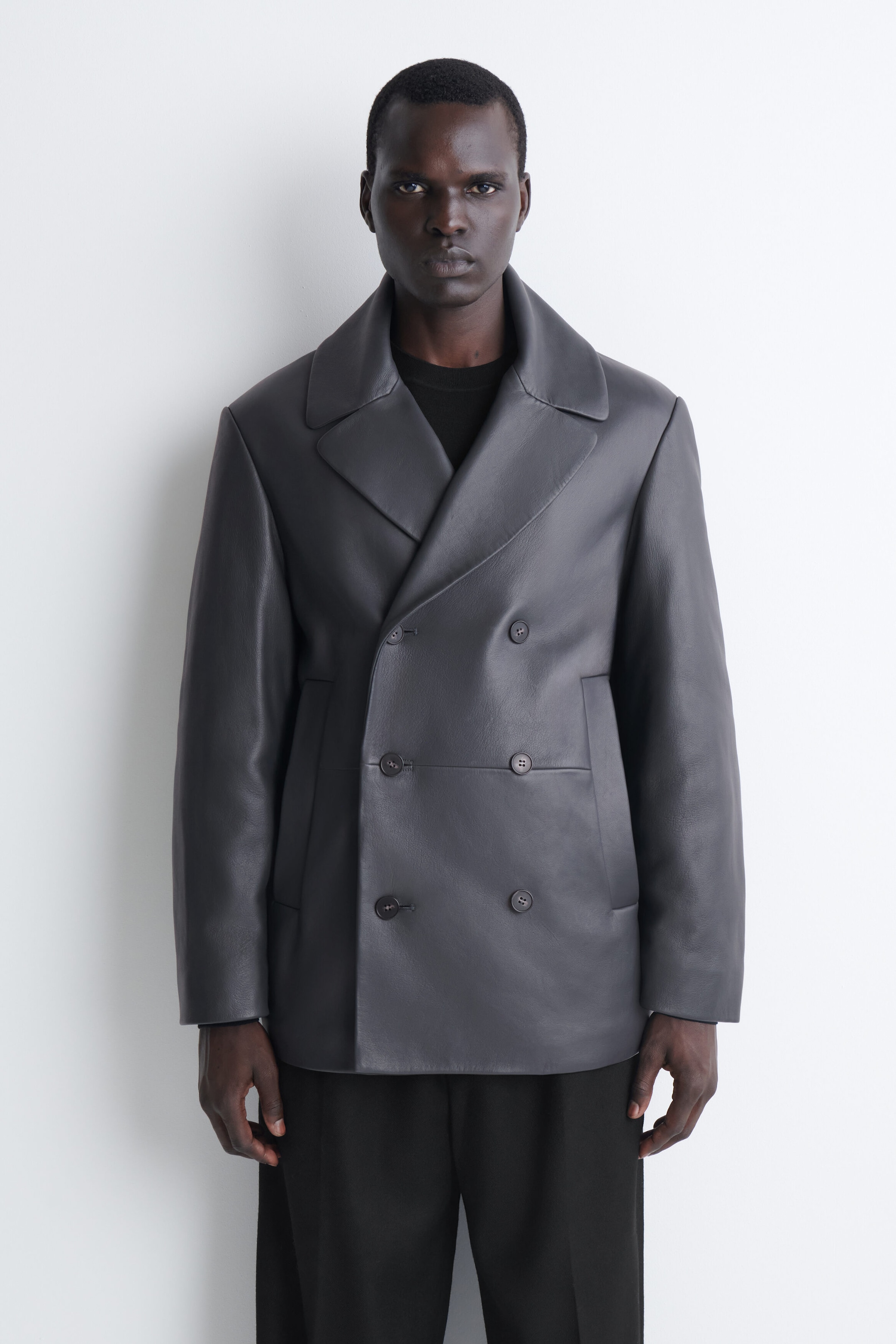 Eine dunkelgraue Lederjacke mit zweireihigem Verschluss ist von vorne zu sehen und verfügt über breite Revers mit fallendem Reversbruch, einen Sechs-Knopf-Verschluss und vertikale Leistentaschen. Die Jacke wird über einem schwarzen Rundhalsshirt und einer schwarzen Hose getragen und betont ihre strukturierte Schulterpartie sowie die glatte, matte Ledertextur. Eine dunkelgraue Lederjacke mit zweireihigem Verschluss ist von vorne zu sehen und verfügt über breite Revers mit fallendem Reversbruch, einen Sechs-Knopf-Verschluss und vertikale Leistentaschen. Die Jacke wird über einem schwarzen Rundhalsshirt und einer schwarzen Hose getragen und betont ihre strukturierte Schulterpartie sowie die glatte, matte Ledertextur.