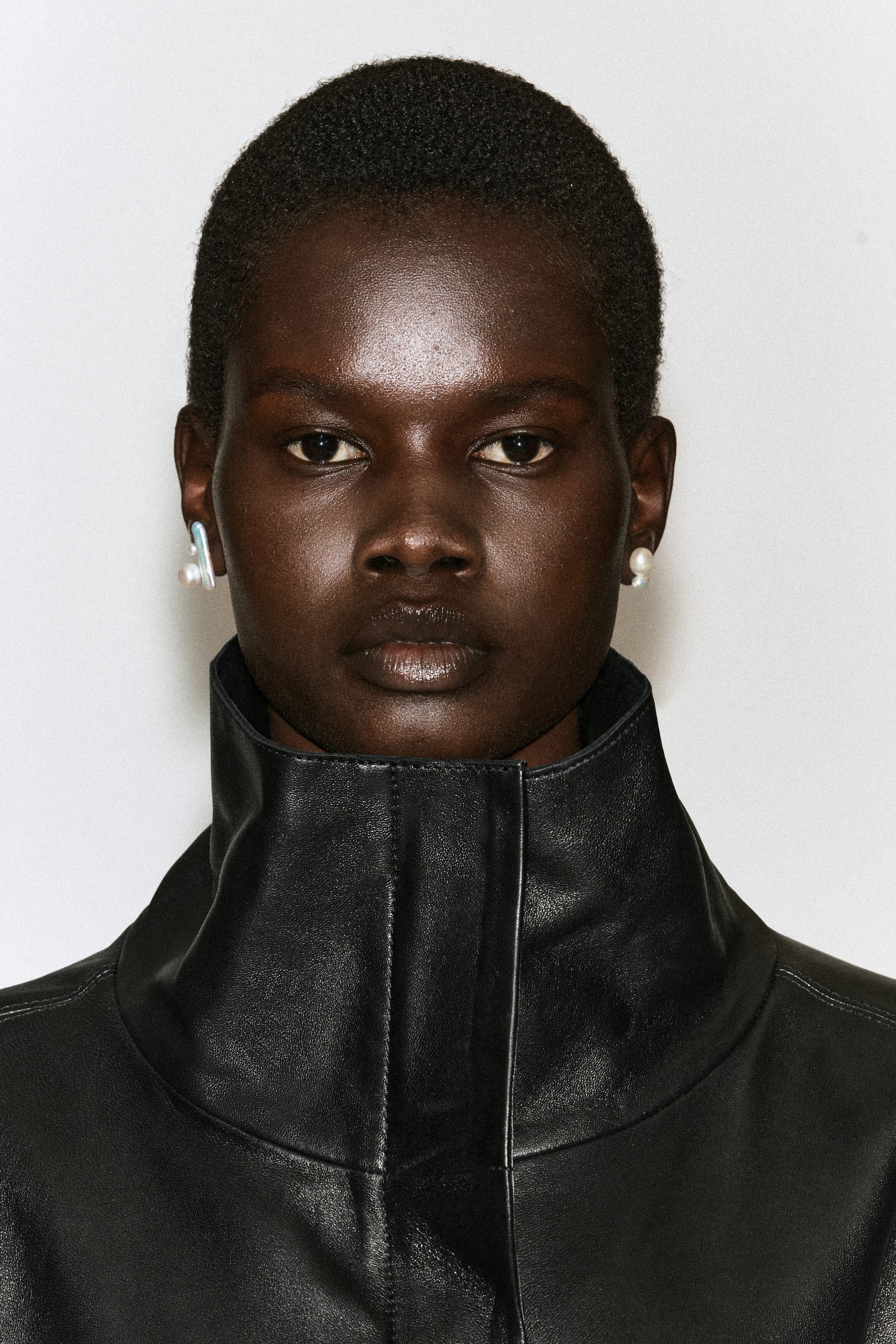 The model is wearing sculptural freshwater pearl earrings, featuring a mismatched pair with an elongated, iridescent bar-shaped pearl on one side and a classic round pearl on the other. The jewellery is styled with a high-collared black leather jacket that frames the face. The model is wearing sculptural freshwater pearl earrings, featuring a mismatched pair with an elongated, iridescent bar-shaped pearl on one side and a classic round pearl on the other. The jewellery is styled with a high-collared black leather jacket that frames the face.