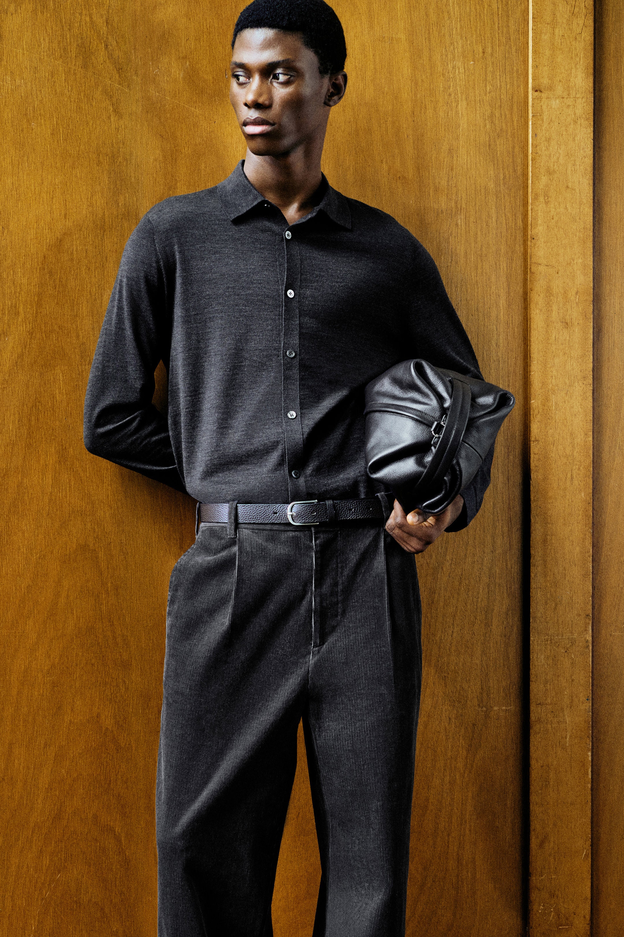 A charcoal-grey knitted merino wool shirt is shown from the front, featuring a classic pointed collar, a button-front fastening with contrasting light buttons, and long sleeves. The shirt drapes softly and is tucked into belted trousers, highlighting its regular fit and fine-knit texture.