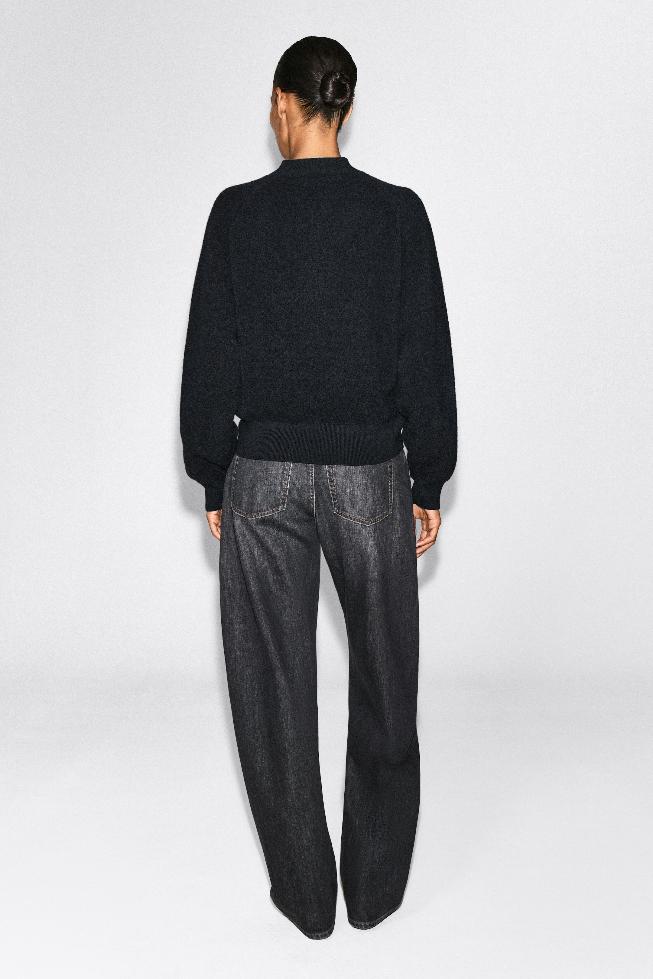 A textured black knit sweater with raglan sleeves and a ribbed hem is worn by the model, styled with dark gray, wide-leg denim jeans. A textured black knit sweater with raglan sleeves and a ribbed hem is worn by the model, styled with dark gray, wide-leg denim jeans.