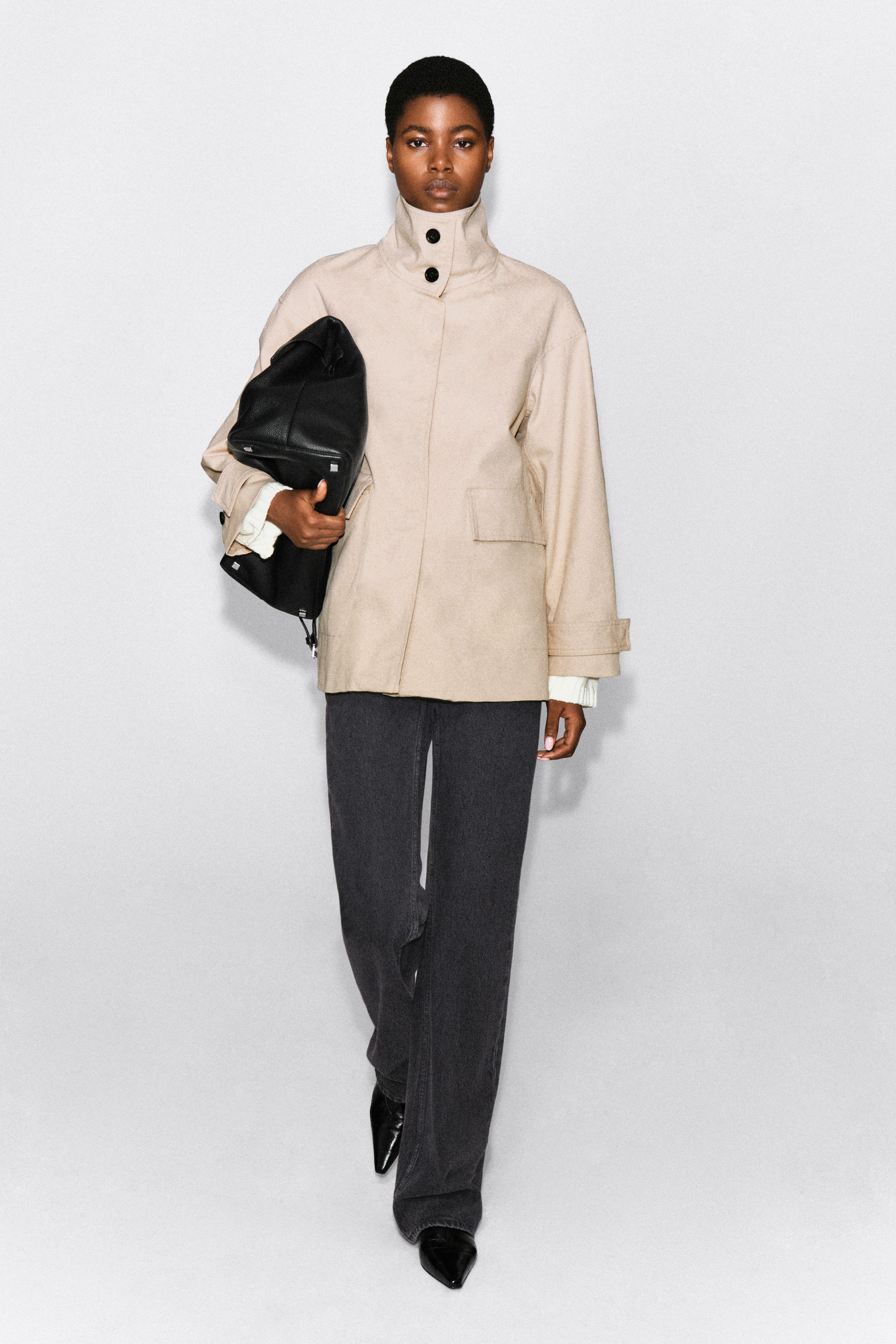 Ver imagen más grande: Una chaqueta de sarga de algodón beige con cuello chimenea y un cuello alto con dos botones negros la lleva puesta la modelo. La chaqueta se combina con pantalones anchos de color gris oscuro, y se lleva bajo el brazo un gran bolso de cuero negro.
