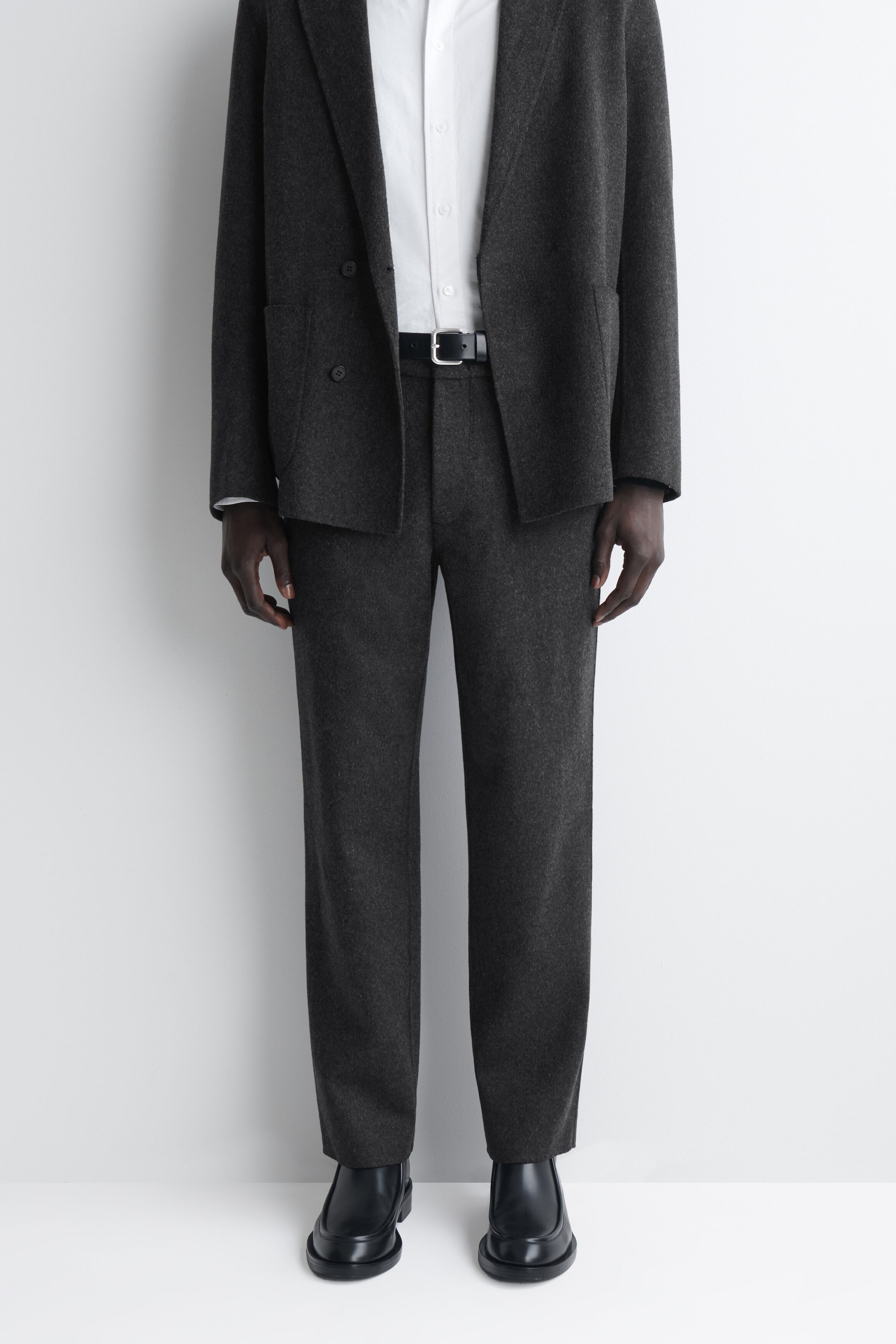 These dark grey, straight-leg trousers are worn in a front view, showcasing their relaxed fit and unstructured design. They are styled with a black leather belt, a matching dark grey blazer, a white button-down shirt, and black leather shoes. These dark grey, straight-leg trousers are worn in a front view, showcasing their relaxed fit and unstructured design. They are styled with a black leather belt, a matching dark grey blazer, a white button-down shirt, and black leather shoes.