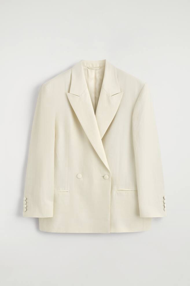 Ein cremefarbener, zweireihiger Smoking-Blazer ist von vorne zu sehen und zeichnet sich durch breite, leicht glänzende Notch-Revers, eine gerade Silhouette und definierte Schultern aus. Er präsentiert vier runde, bezogene Knöpfe am Verschluss vorne, drei bezogene Knöpfe an jedem Ärmelabschluss und zwei Pattentaschen an den Hüften. - 1