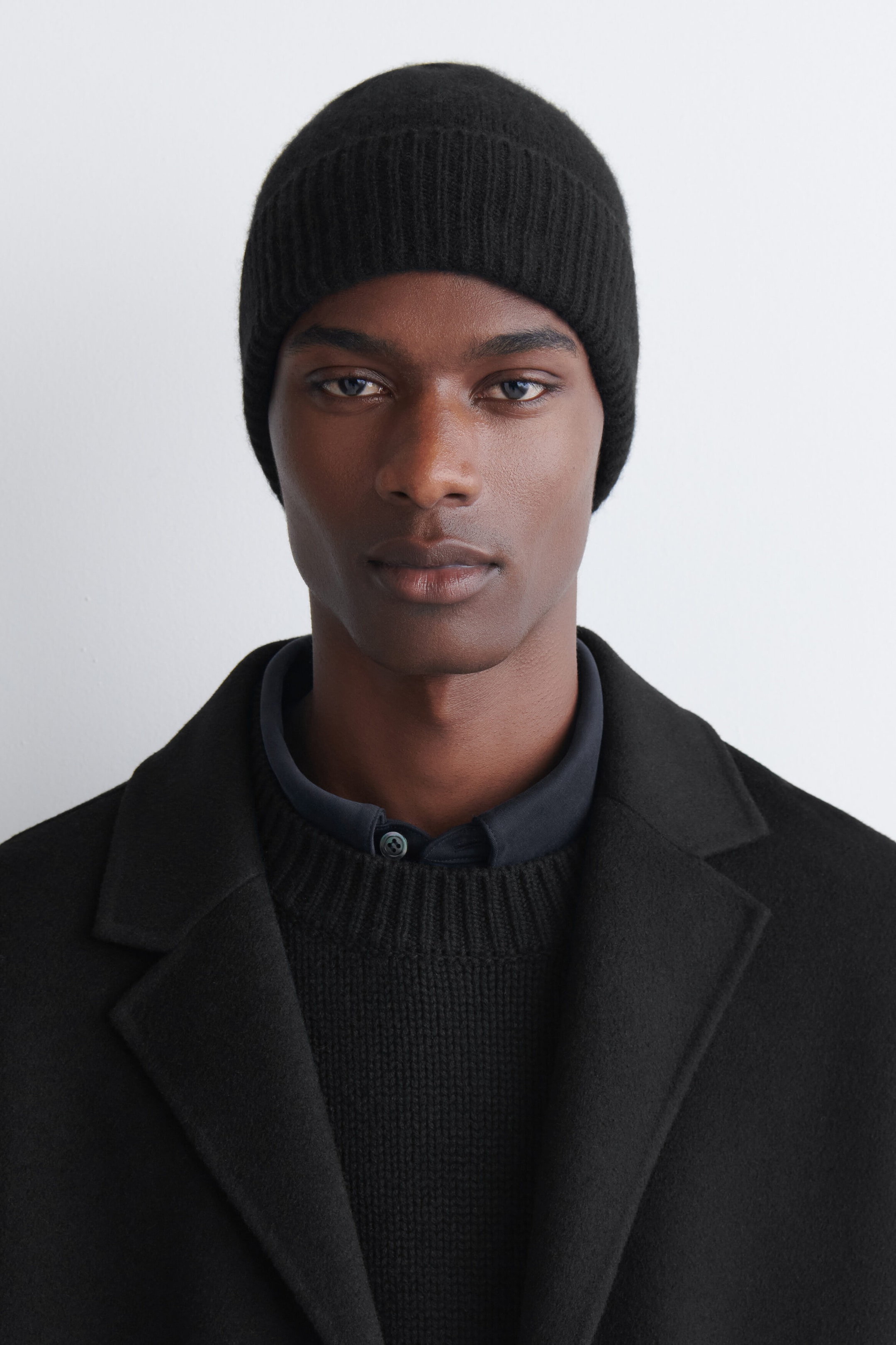 A black cashmere beanie featuring a ribbed-knit construction and a wide folded cuff is being worn facing front. The beanie fits snugly on the head, highlighting its soft texture and clean design.