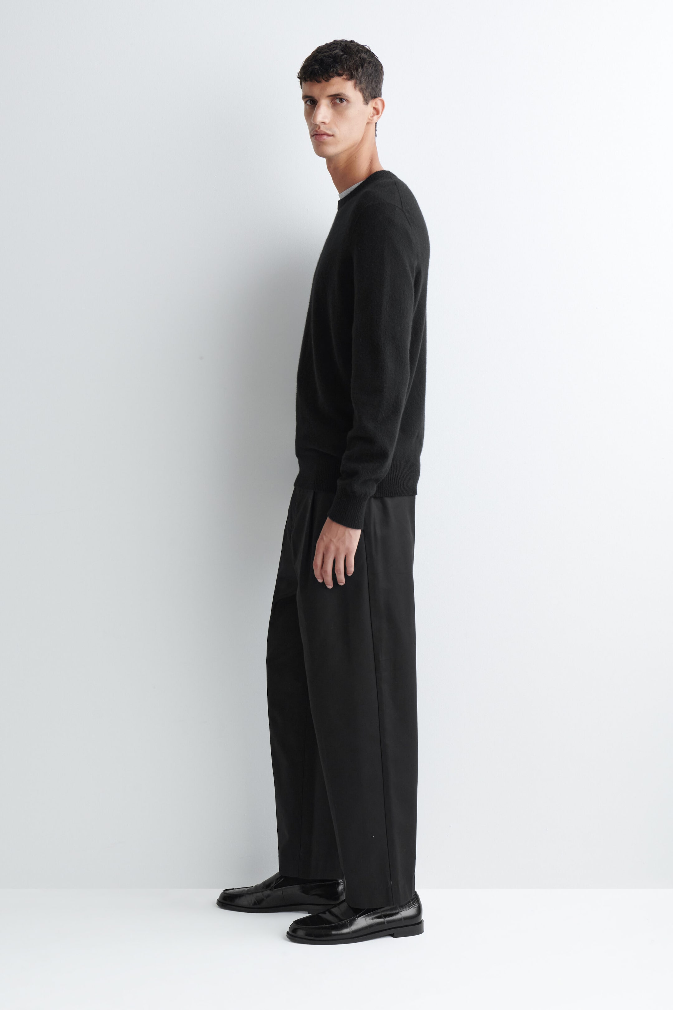 Das Model trägt einen schwarzen Pullover mit Rundhalsausschnitt und langen Ärmeln, der an Bündchen und Saum mit Rippdetails versehen ist. Der Pullover ist mit einer schwarzen Wide-Leg-Hose und schwarzen Lackleder-Loafern kombiniert. Das Model trägt einen schwarzen Pullover mit Rundhalsausschnitt und langen Ärmeln, der an Bündchen und Saum mit Rippdetails versehen ist. Der Pullover ist mit einer schwarzen Wide-Leg-Hose und schwarzen Lackleder-Loafern kombiniert.
