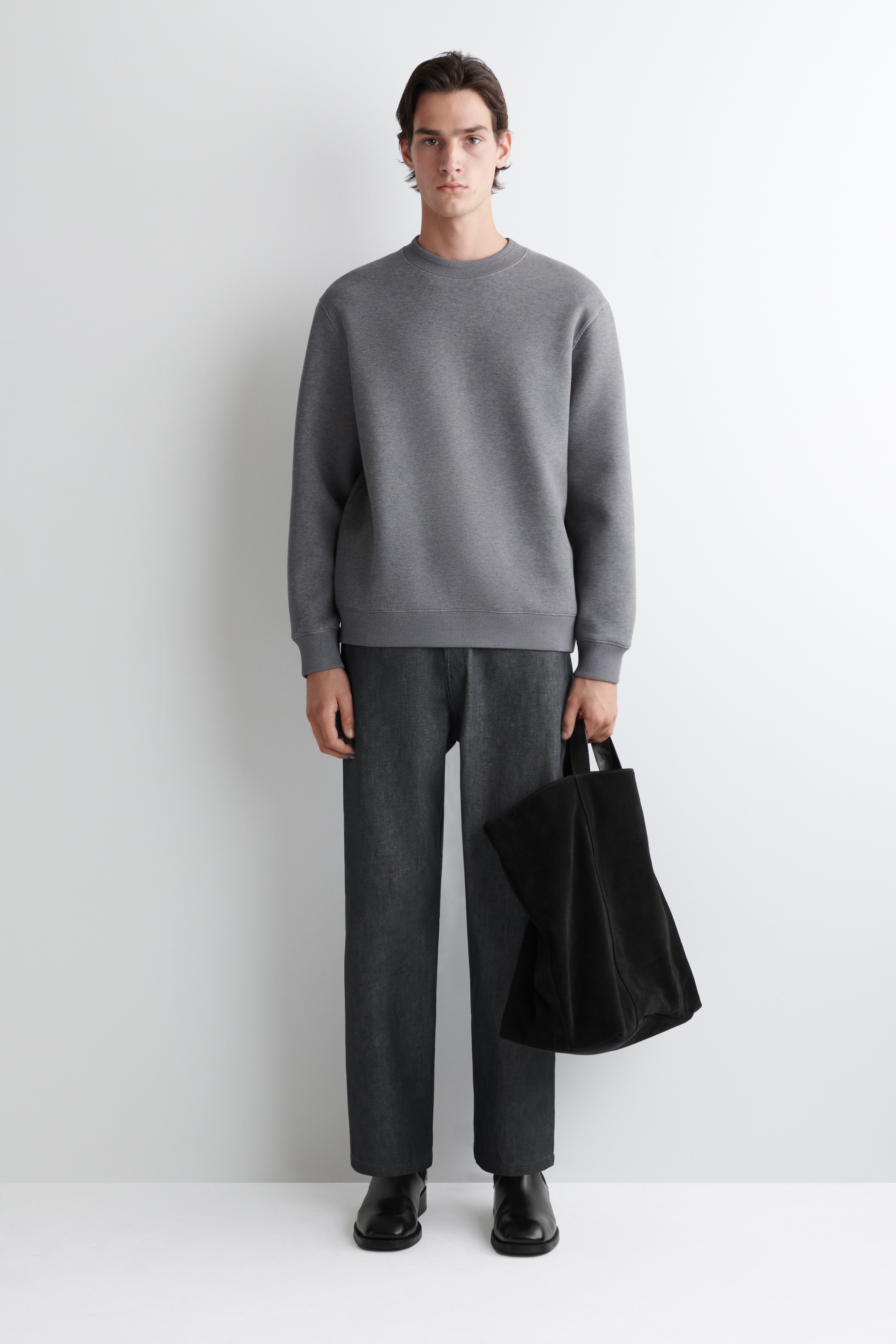 Visualizza immagine più grande: Una felpa girocollo grigio carbone con polsini a coste è indossata con pantaloni grigio scuro a gamba dritta e stivaletti neri a punta quadrata, presentando una vista frontale dell'insieme. Una borsa tote nera non strutturata è tenuta nella mano destra.